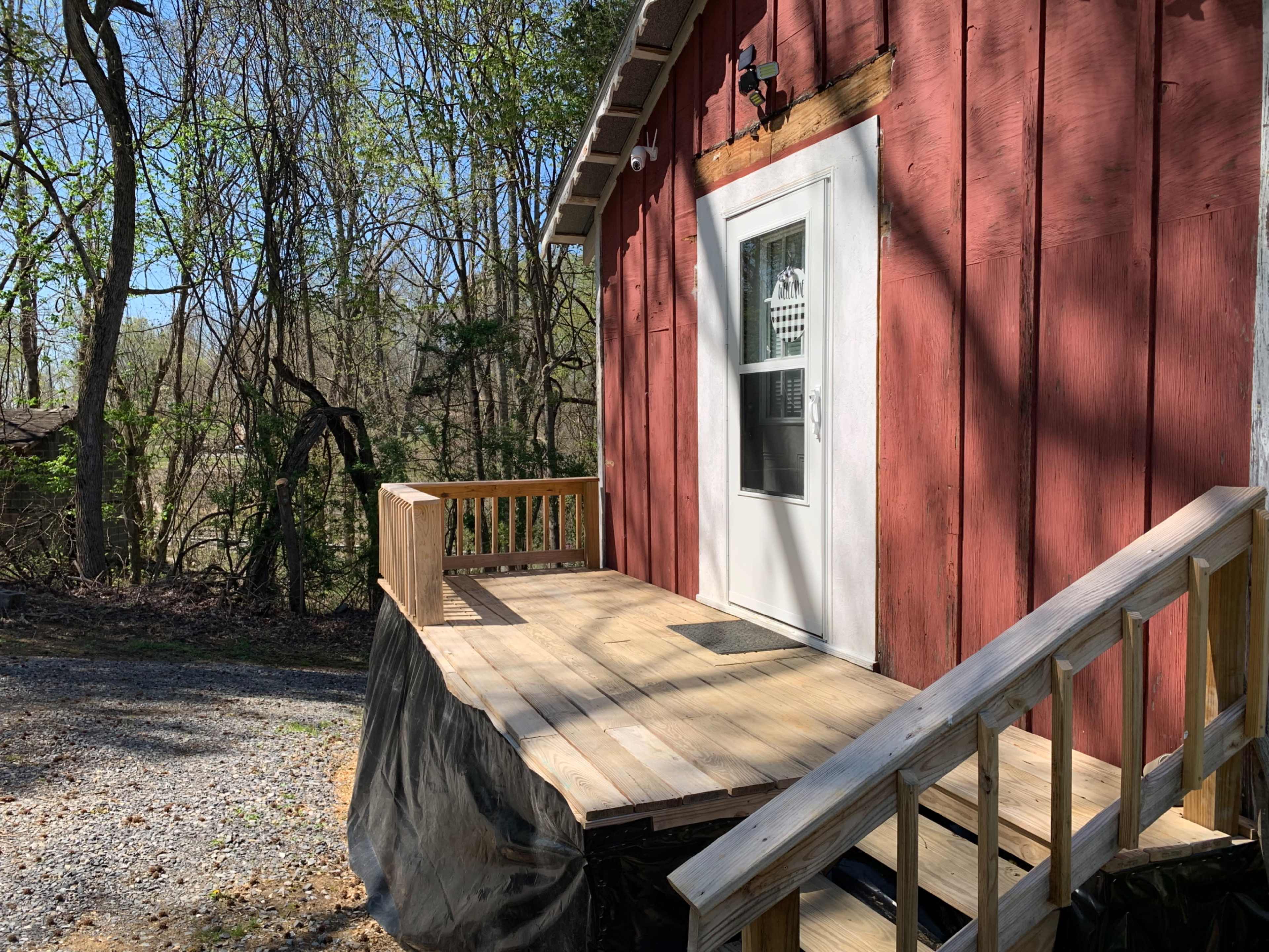 A red wooden cabin features a front porch with a wooden railing and steps leading to a white door.
