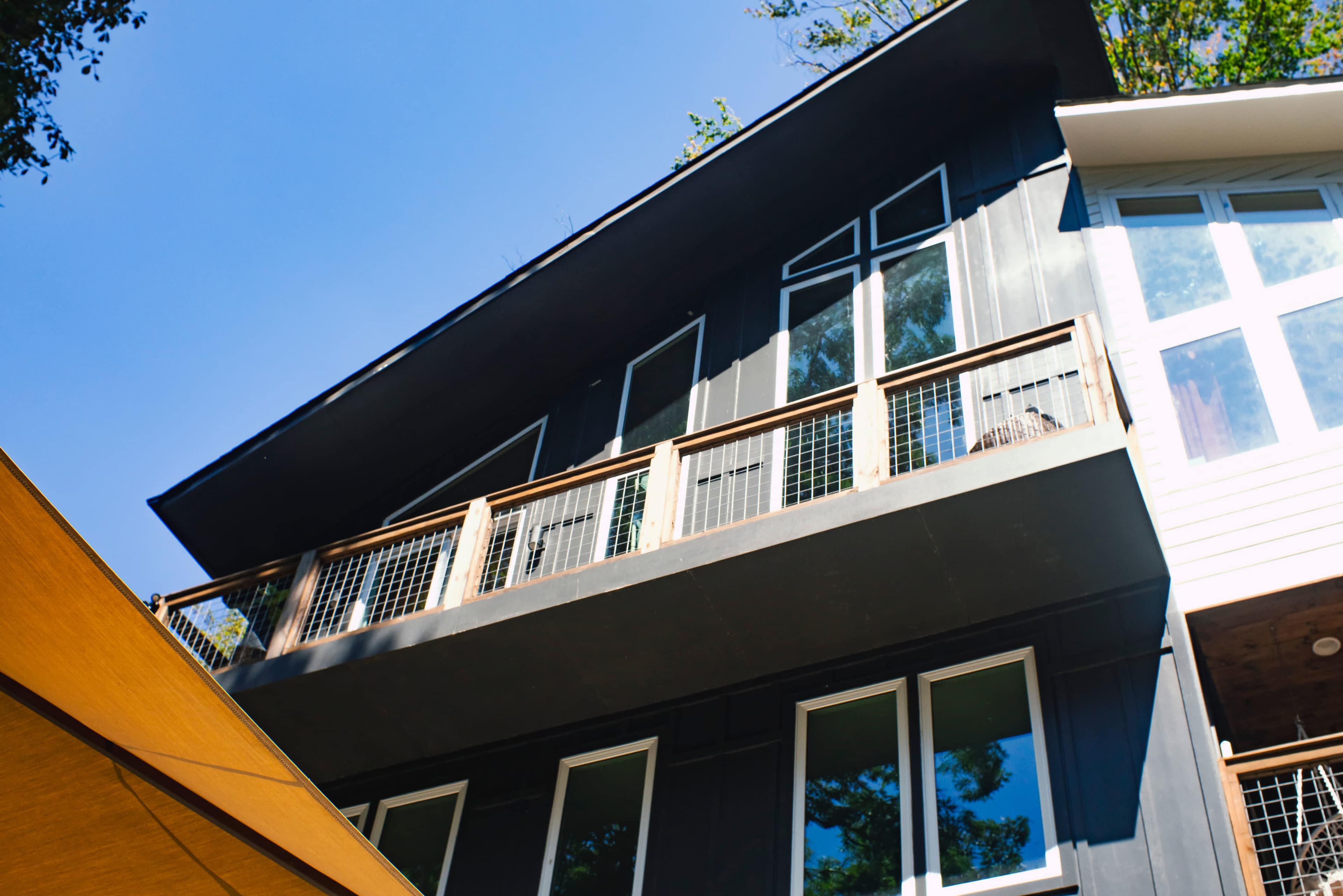 The image shows a modern house with a sloped roof, featuring large windows and a balcony, set against a clear blue sky.
