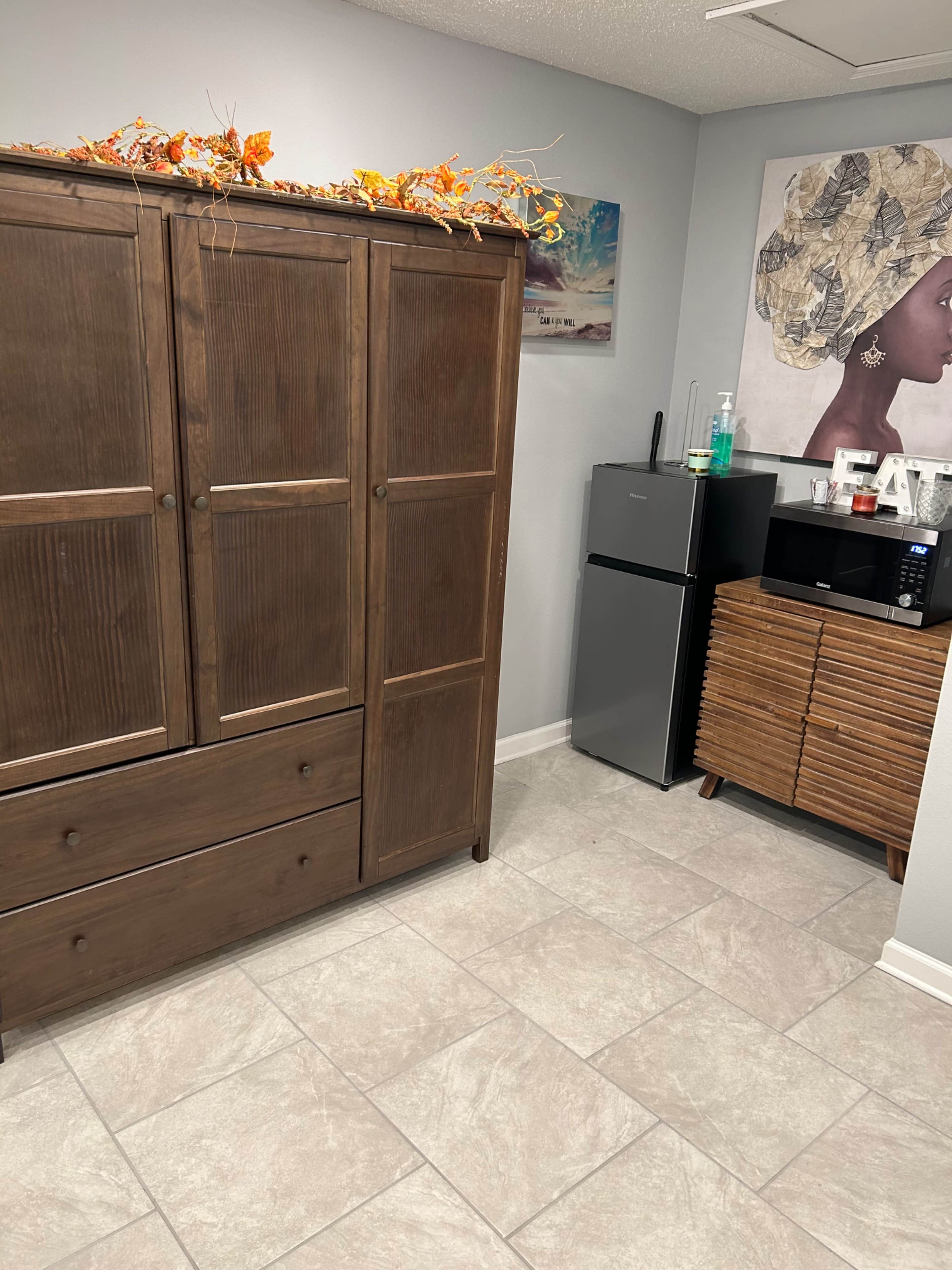 A room with a wooden wardrobe, a small fridge, a microwave, and patterned décor on the wall.