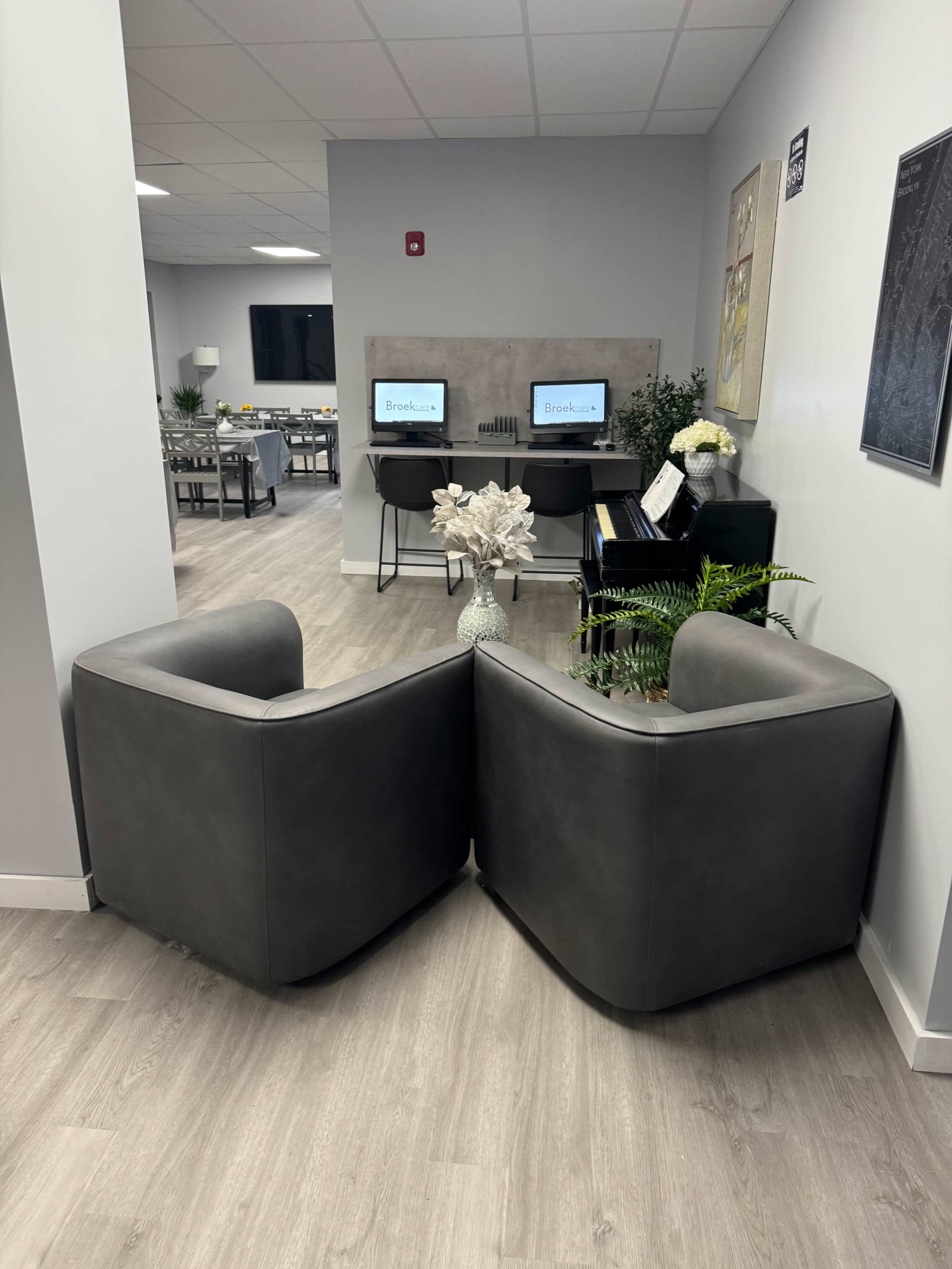 The image shows a modern seating area with two gray chairs facing each other, separated by a small decorative table, and a workspace with two computer stations in the background.