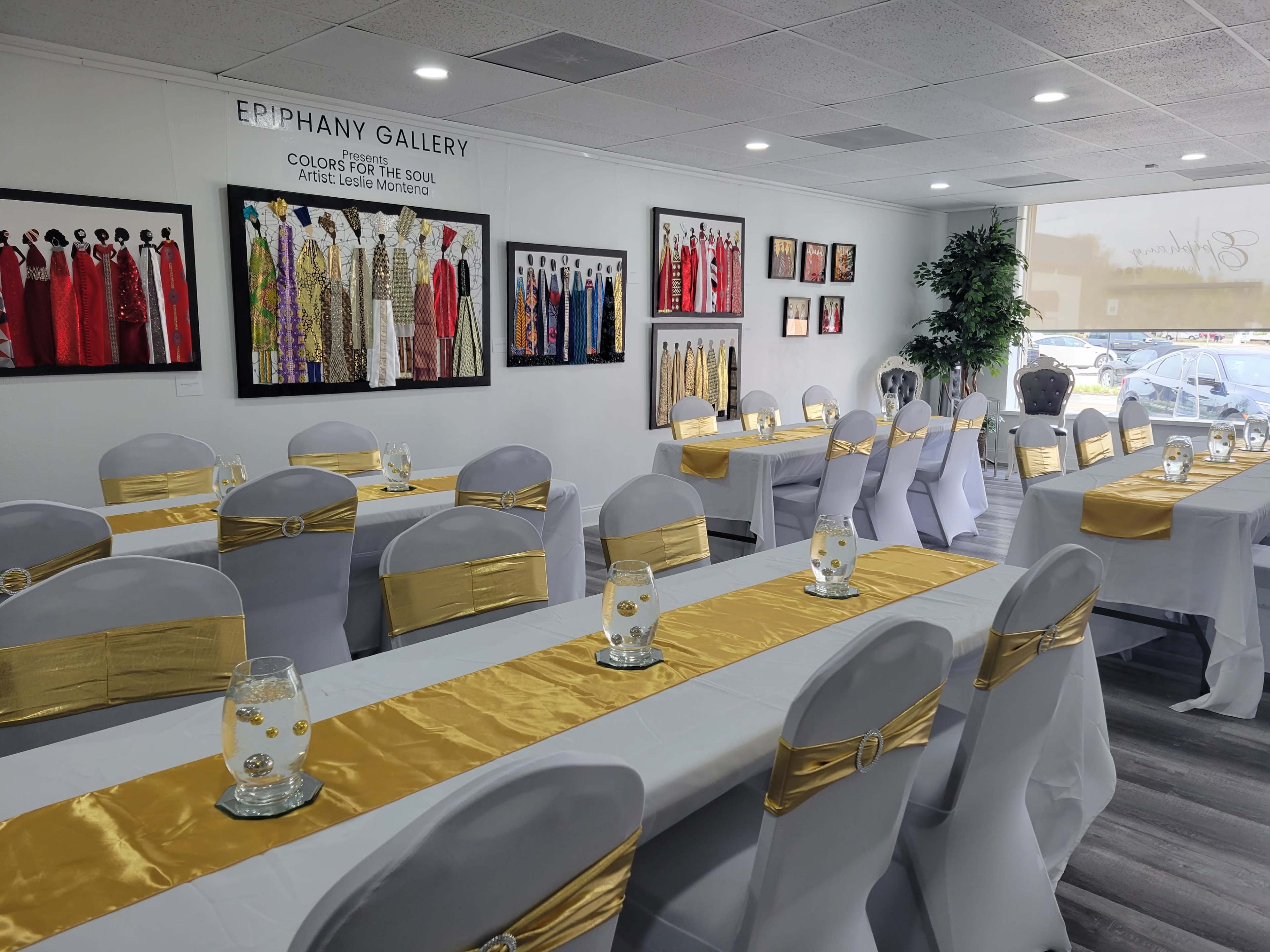 A neatly arranged banquet room with white tables adorned with gold table runners and decorated with glass vases in front of framed artworks on the walls.
