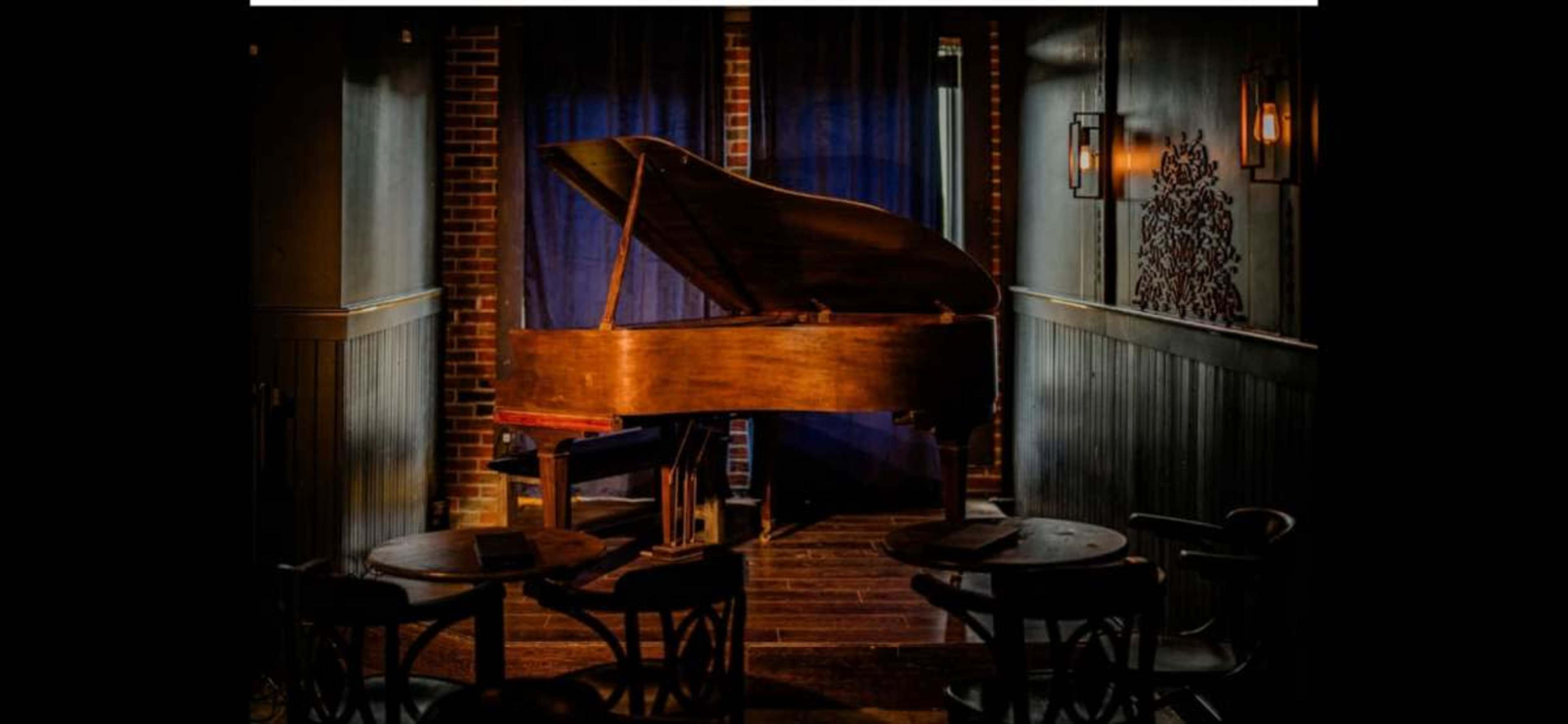 A grand piano sits against a dark blue curtain in a dimly lit room with wooden tables and vintage lighting.