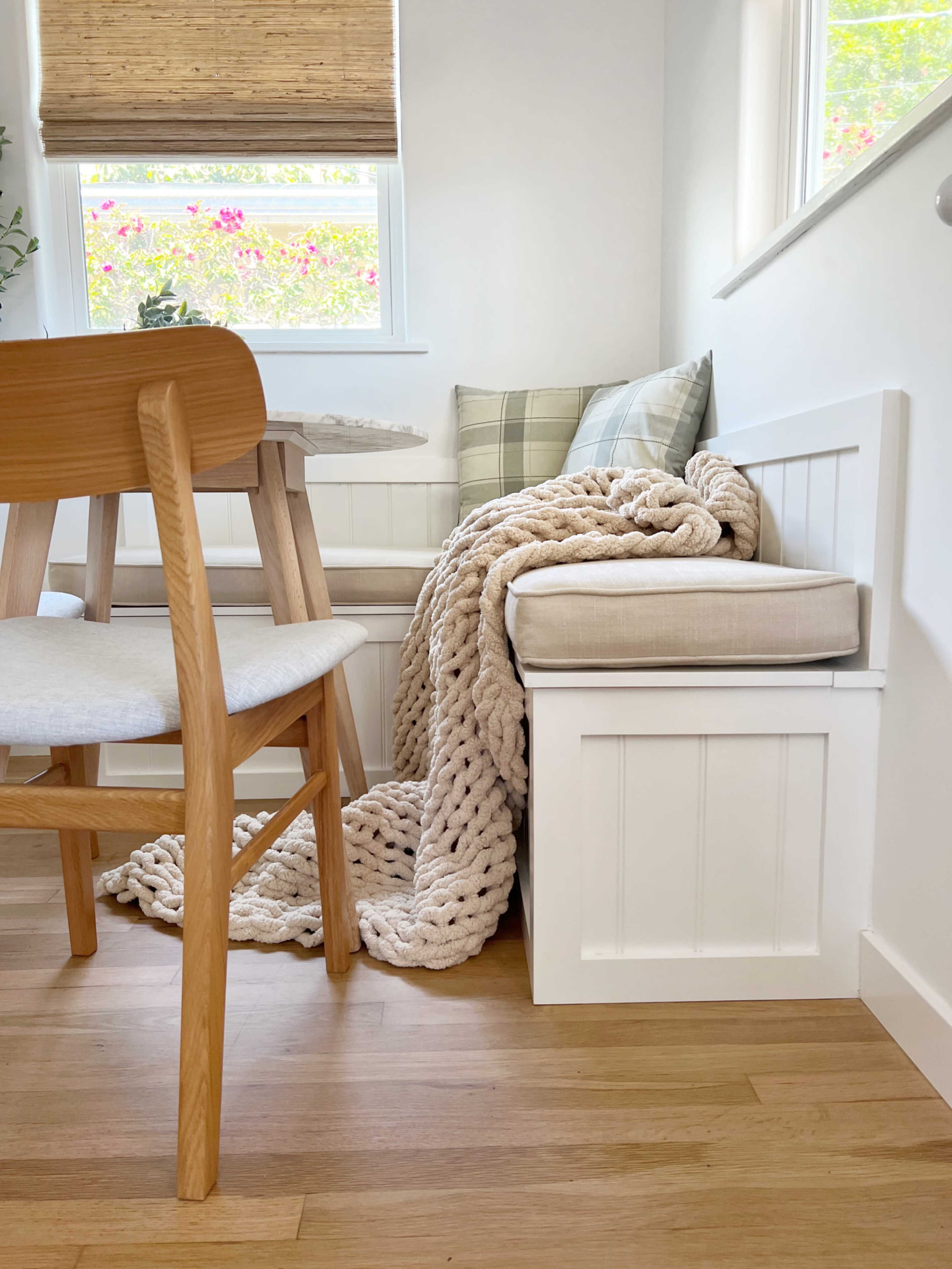 A cozy corner features a wooden chair, a dining table, a cushioned bench with a blanket draped over it, and large windows letting in natural light.