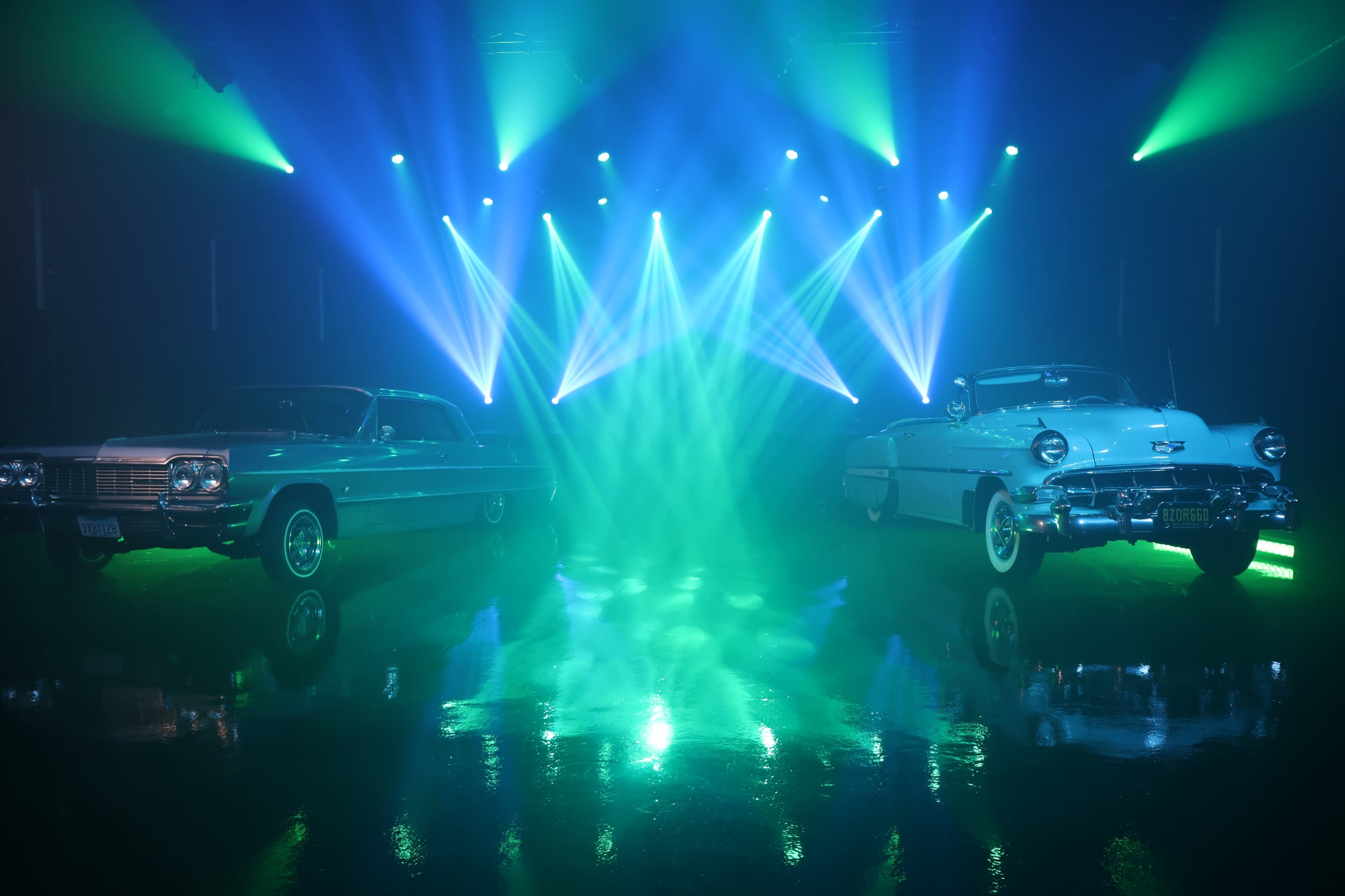 Two classic cars are positioned on a glossy surface, illuminated by vibrant, colored stage lights casting beams in the background.