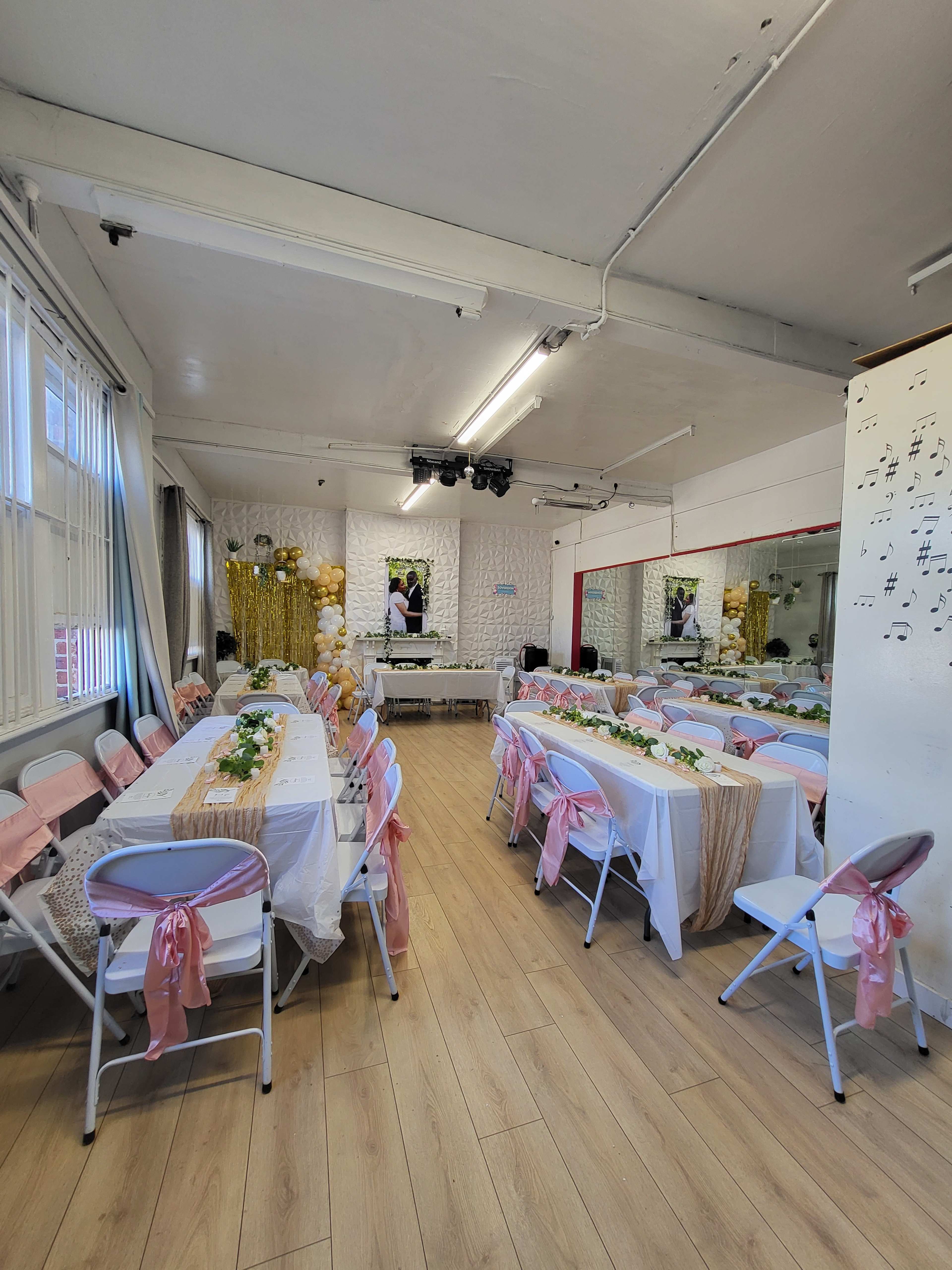 The image shows a decorated event space with tables set for a gathering, featuring white tablecloths and pink sashes on the chairs, along with floral arrangements and a backdrop.