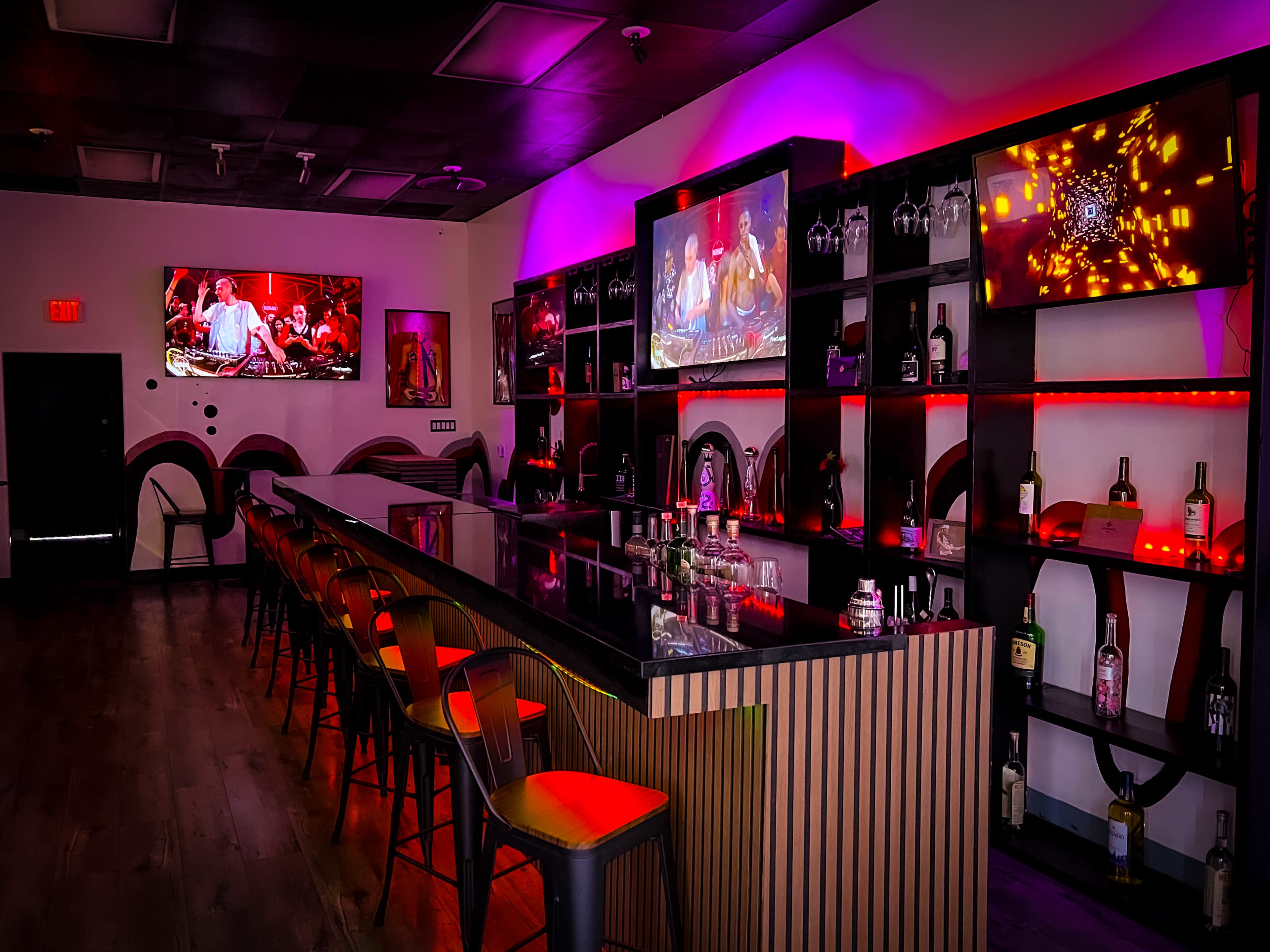 A modern bar area with a polished counter, high stools, and illuminated shelves displaying various bottles, flanked by large screens showing videos.