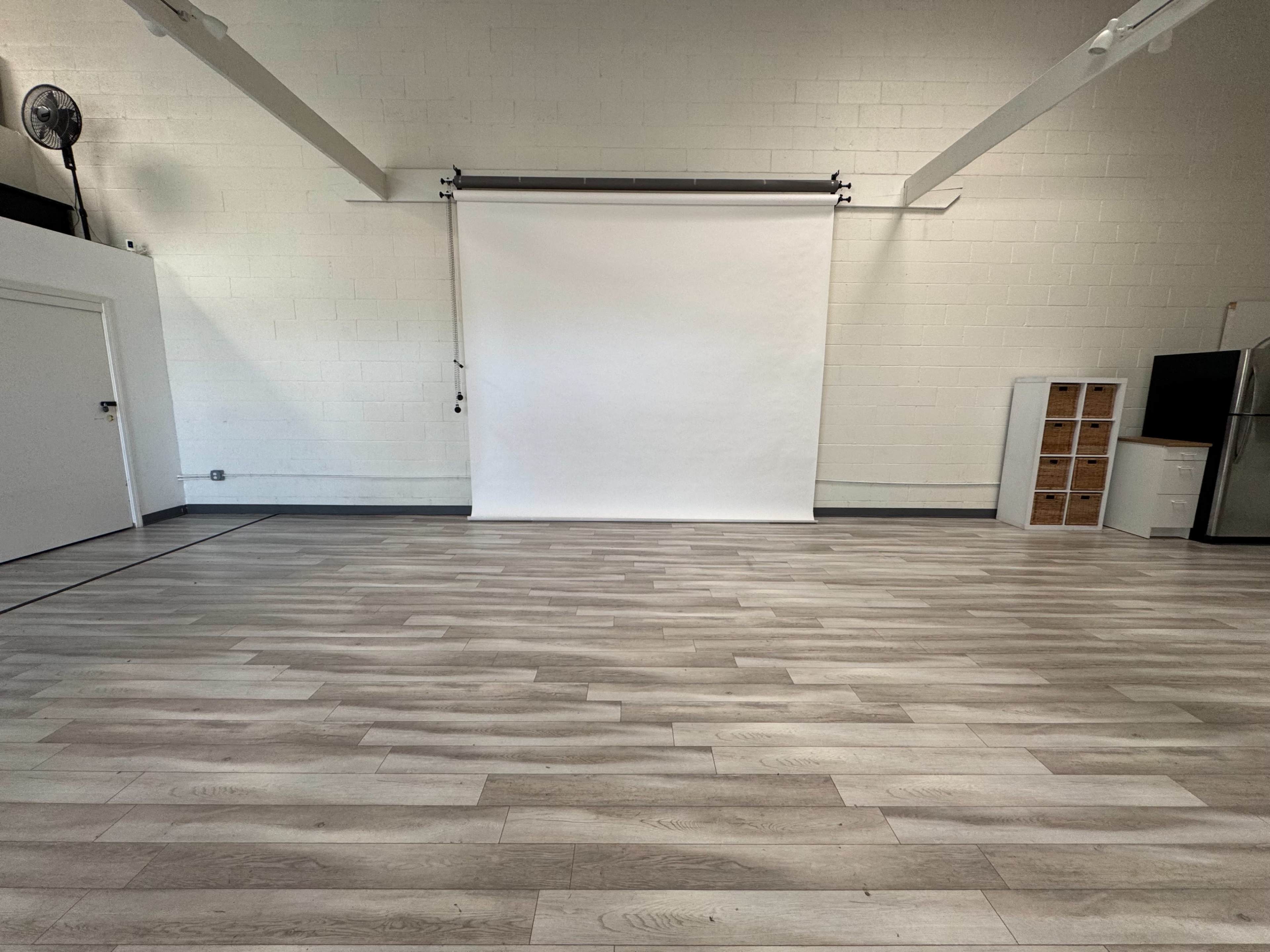 The image shows a spacious, well-lit room with light wooden flooring and a blank white backdrop at one end.