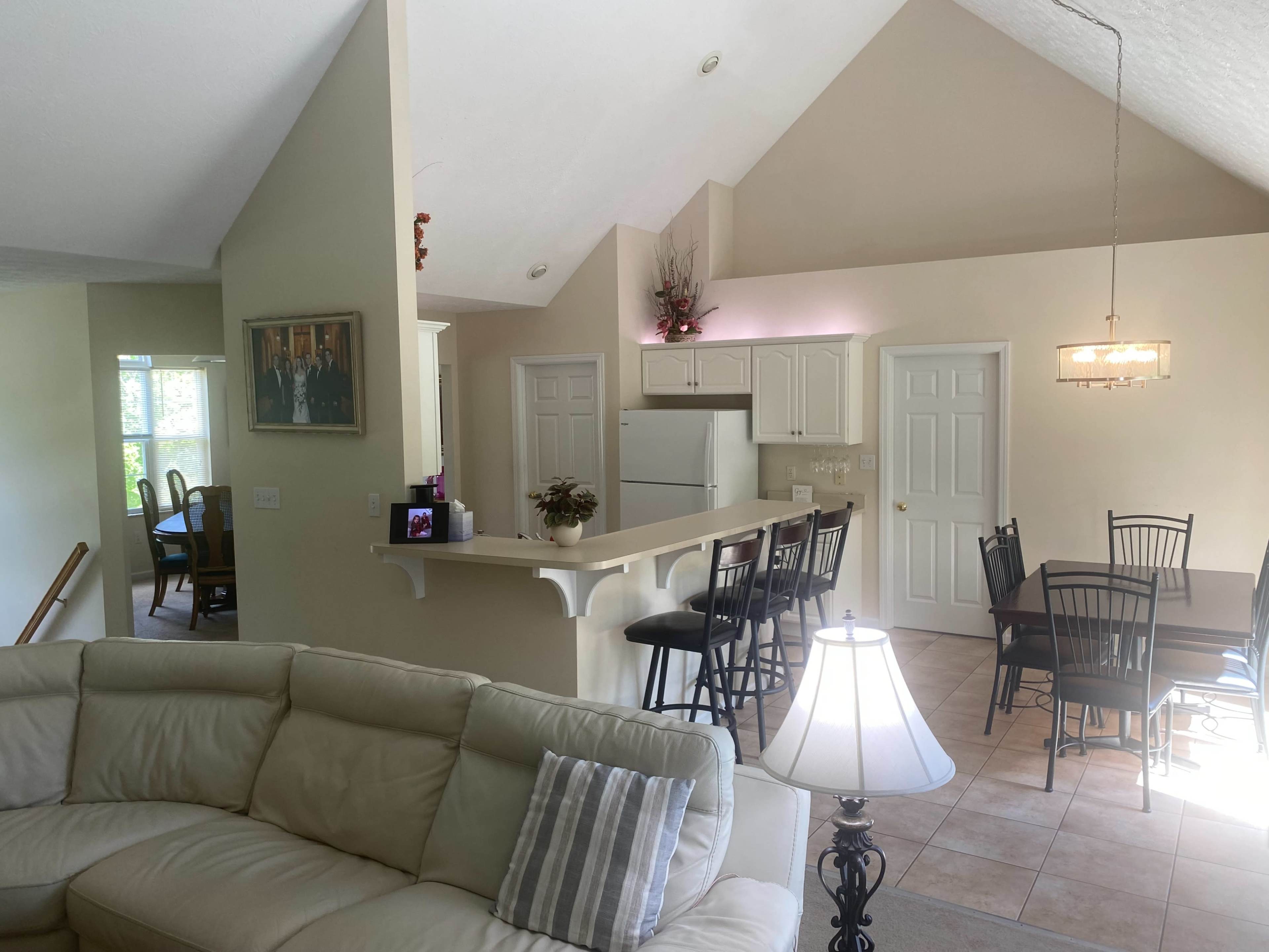 The image shows a bright, open-concept living area with a kitchen and dining space, featuring a light-colored sofa, a bar with stools, and a dining table surrounded by chairs.