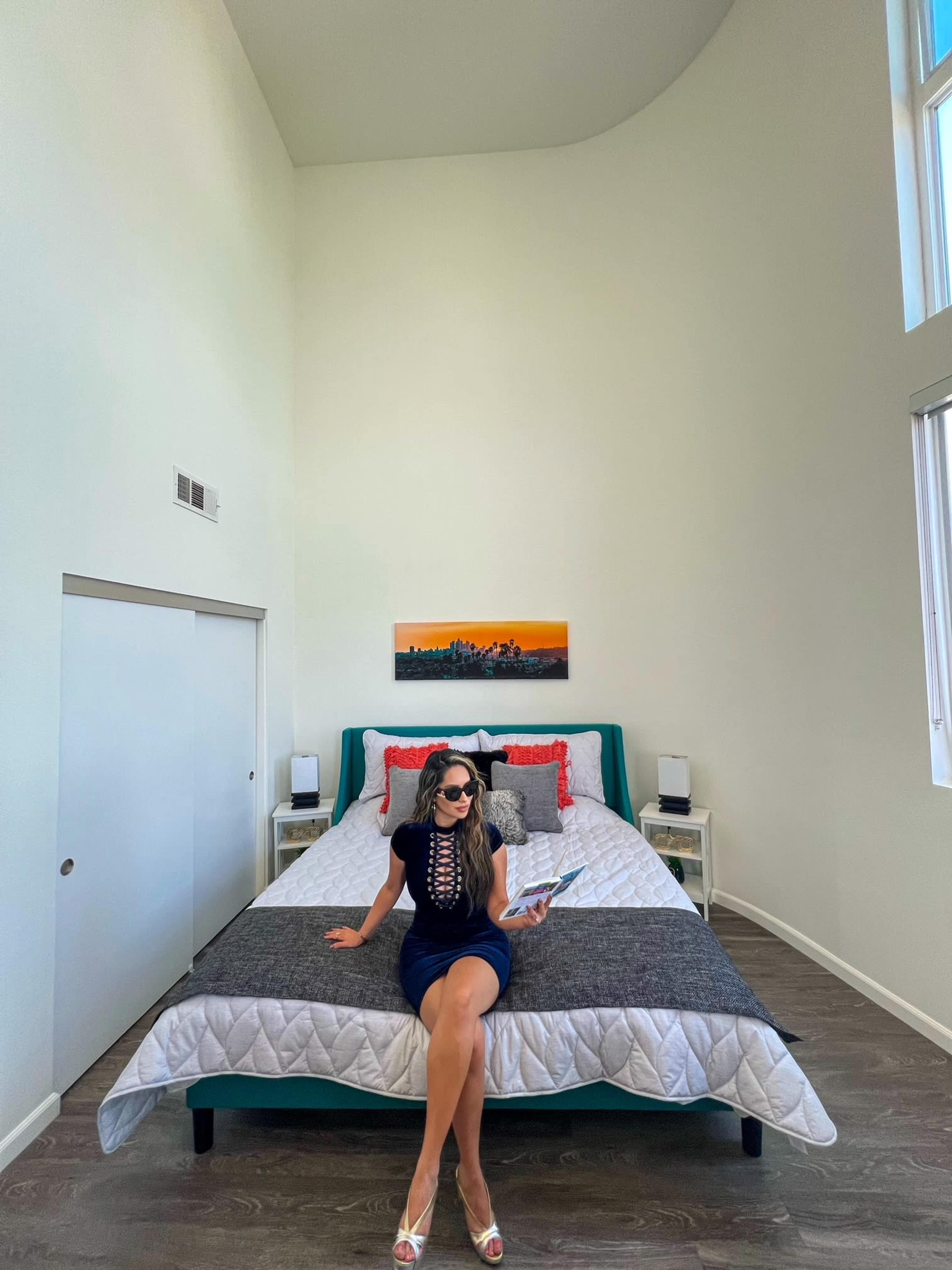 A woman sits on a bed in a brightly lit room with high ceilings, reading a magazine.
