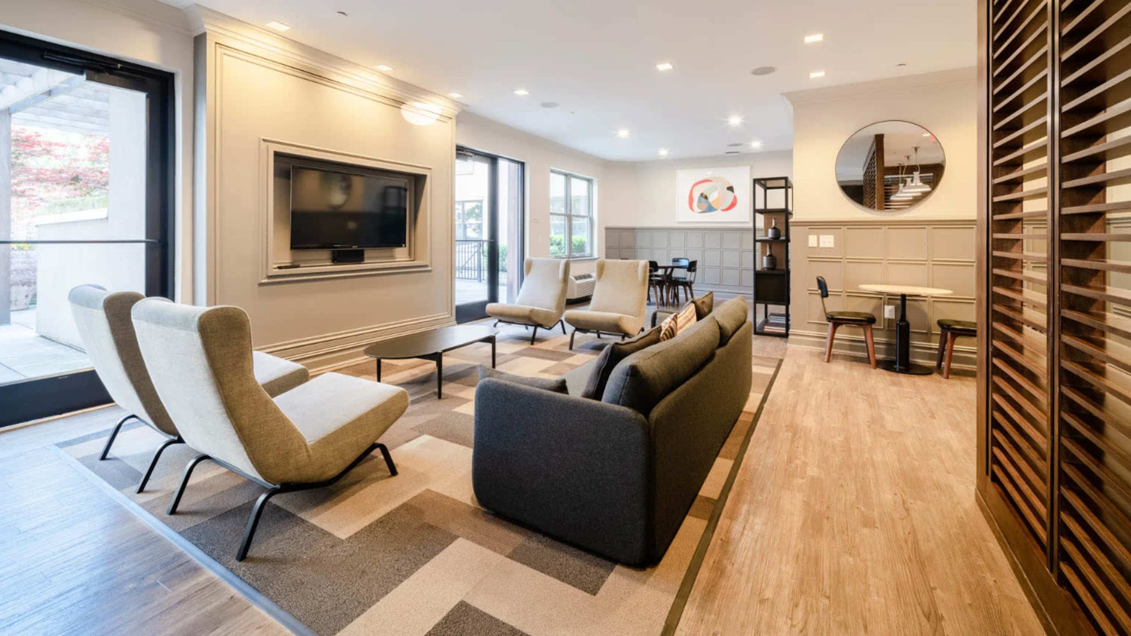 A modern lounge area with seating, a television mounted on the wall, and a small dining space in the background.