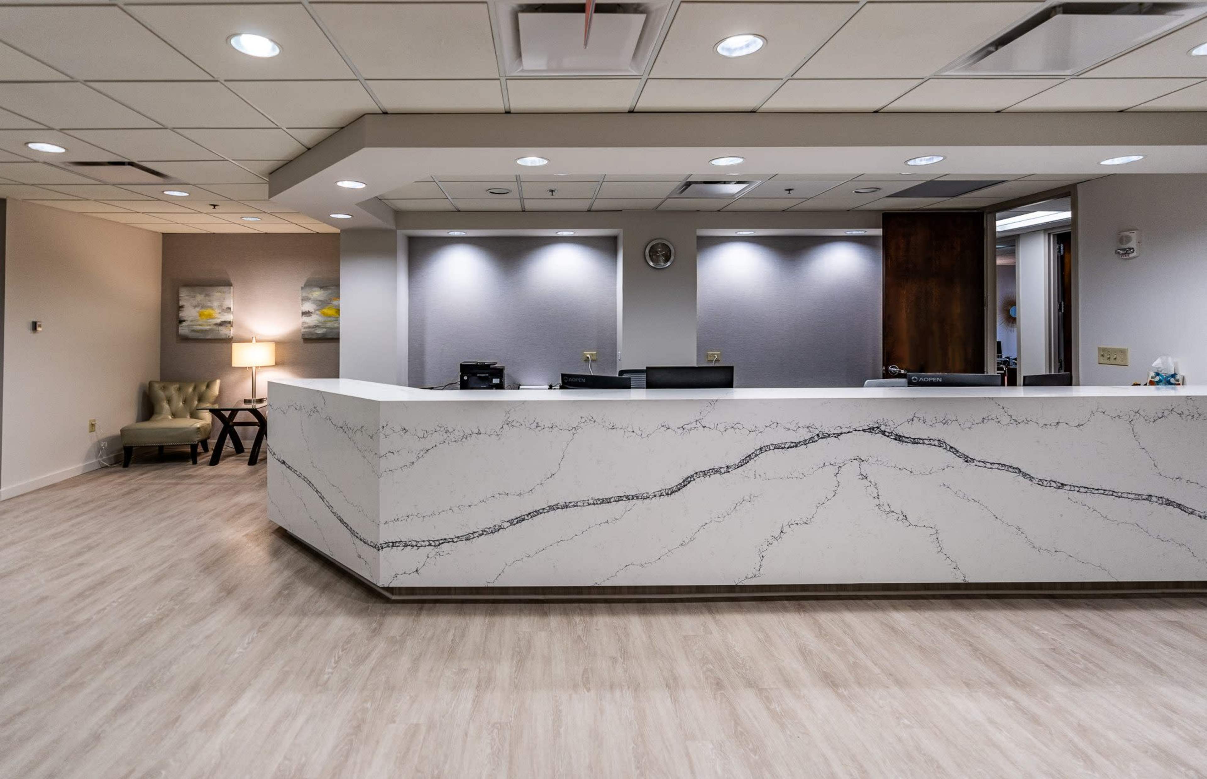 The image shows a modern reception area with a large white marble front desk, wooden accents, and soft lighting.