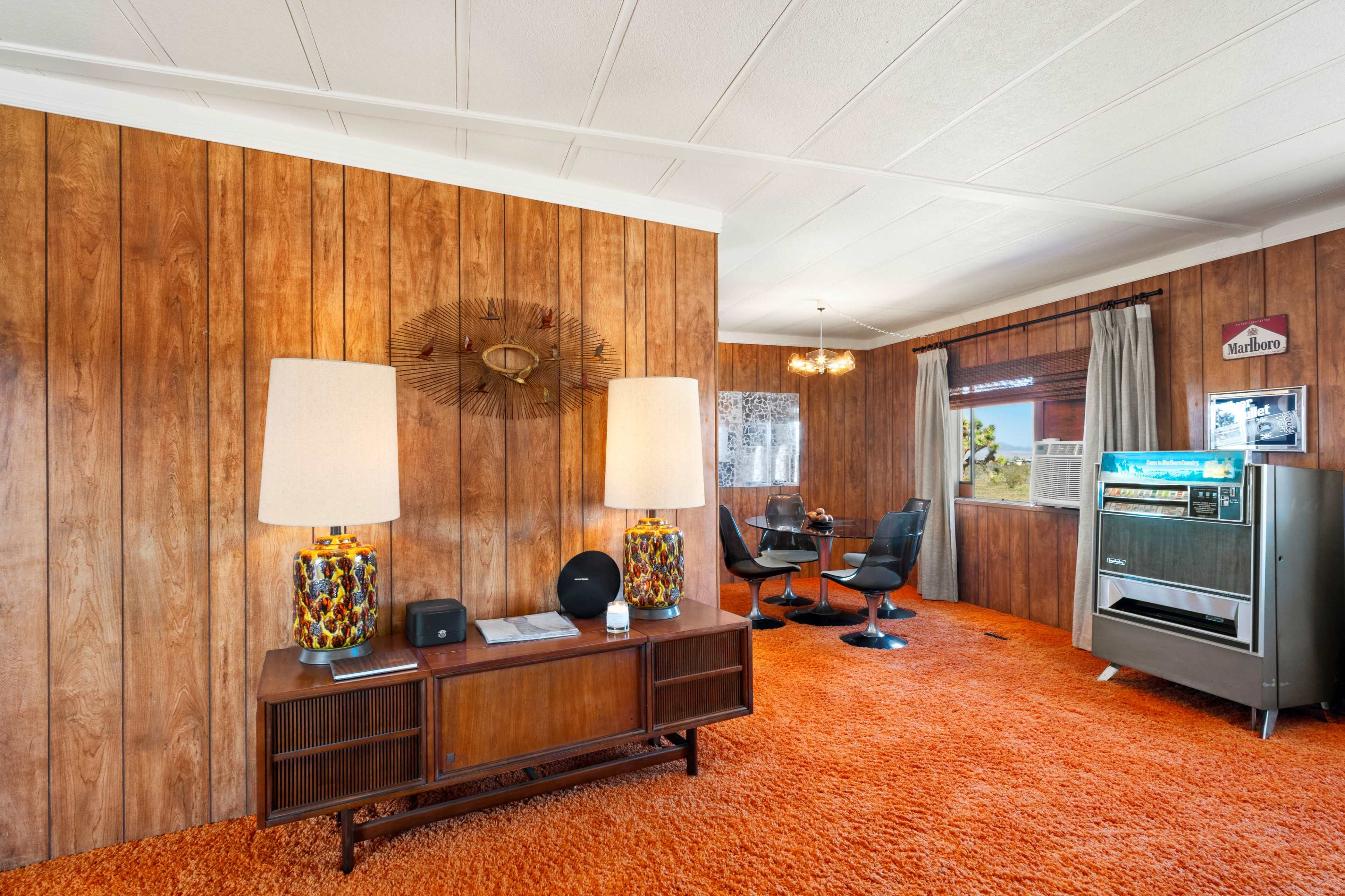 The interior features wooden paneling, orange carpet, two large lamps on a side table, and a dining area with modern chairs and a retro refrigerator.