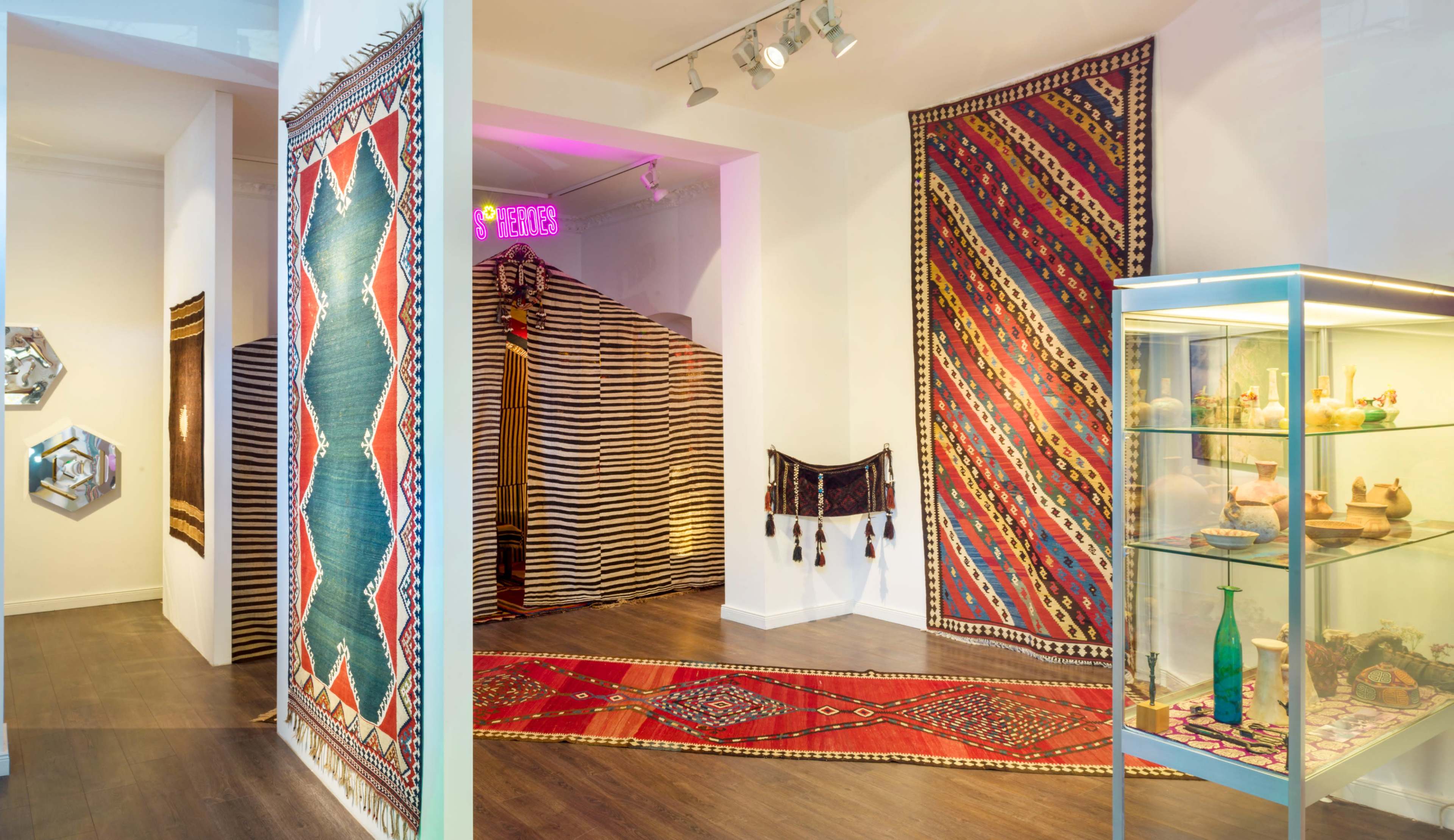 The image shows a gallery space featuring colorful textile artworks displayed on the walls and a glass case containing decorative items.