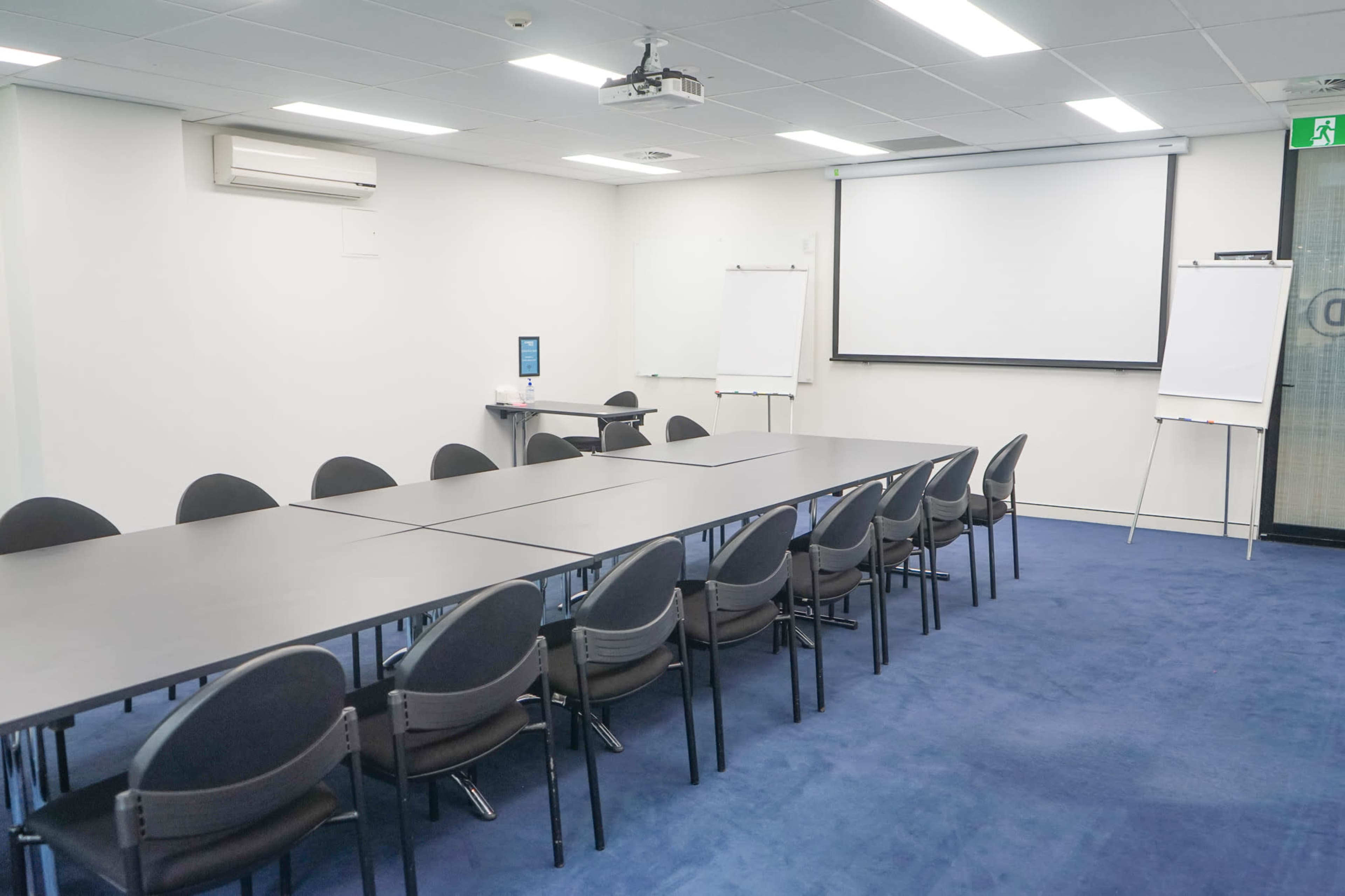 A conference room is set up with a long table and chairs, featuring a projector screen and whiteboards at the front.