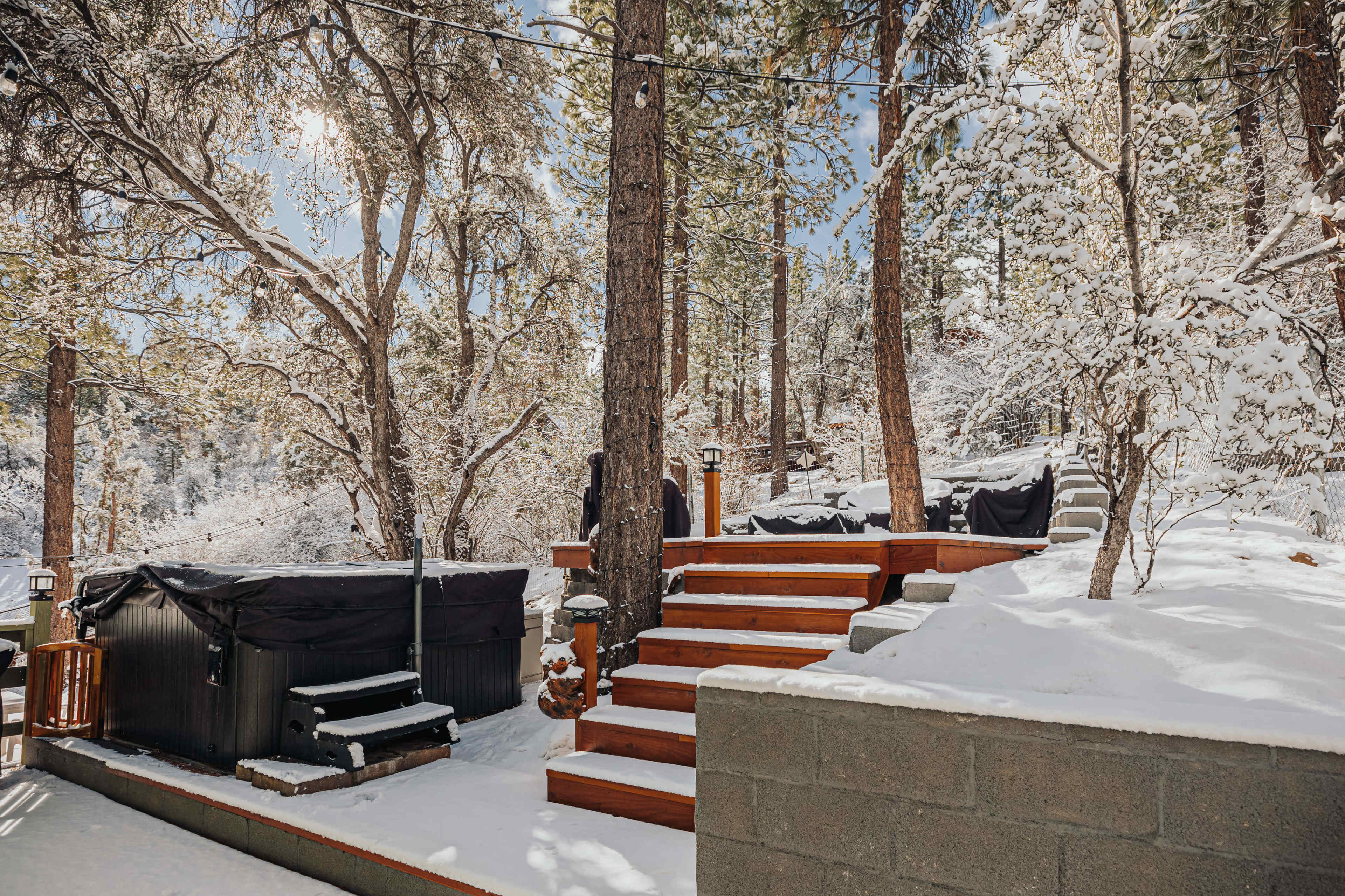 A snowy outdoor scene features steps leading up to a wooden deck, surrounded by pine trees and hot tubs covered with black tarps.