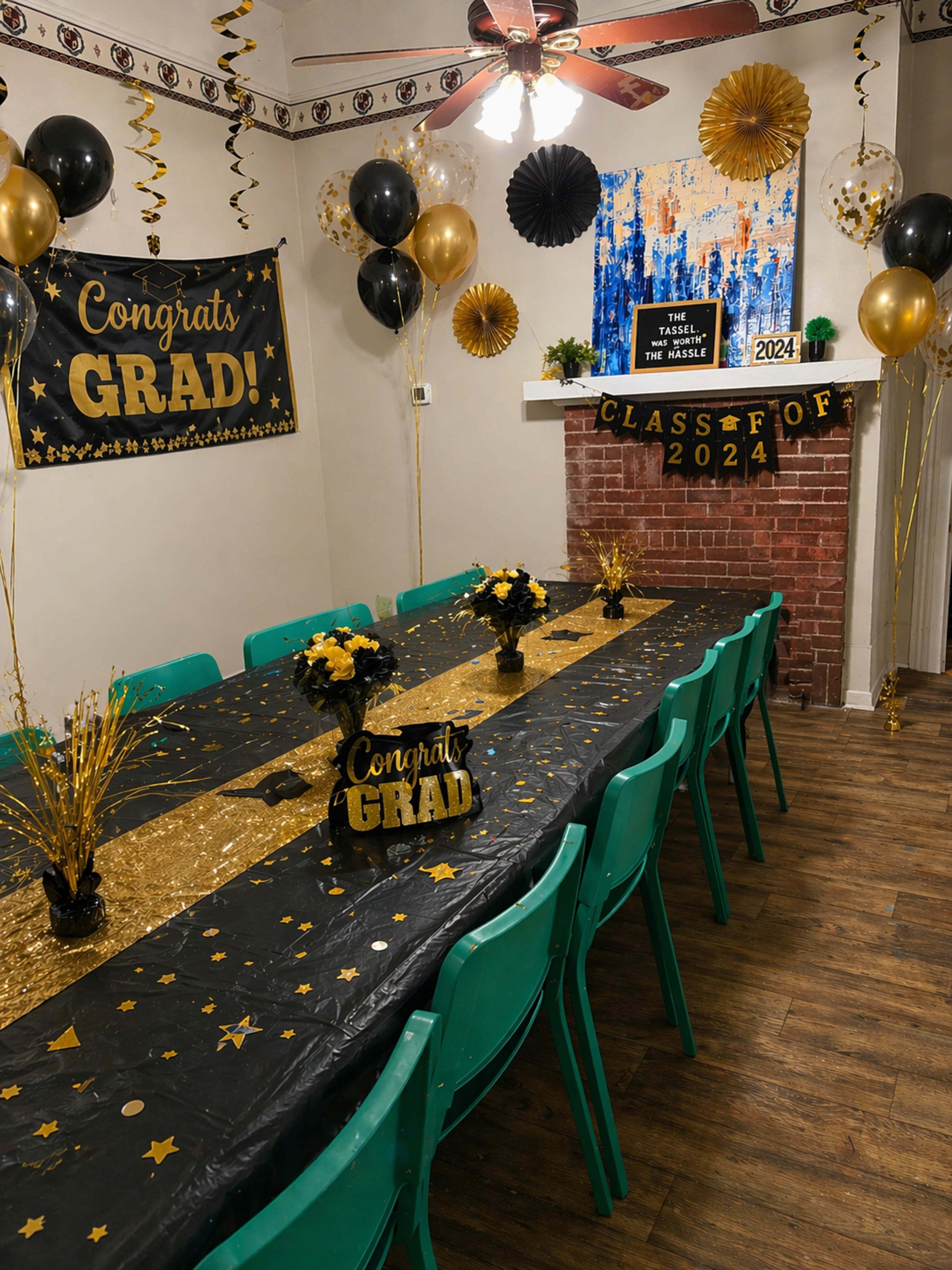 The image shows a decorated room for a graduation celebration, featuring a long table with a black and gold tablecloth, balloons, banners, and floral arrangements.