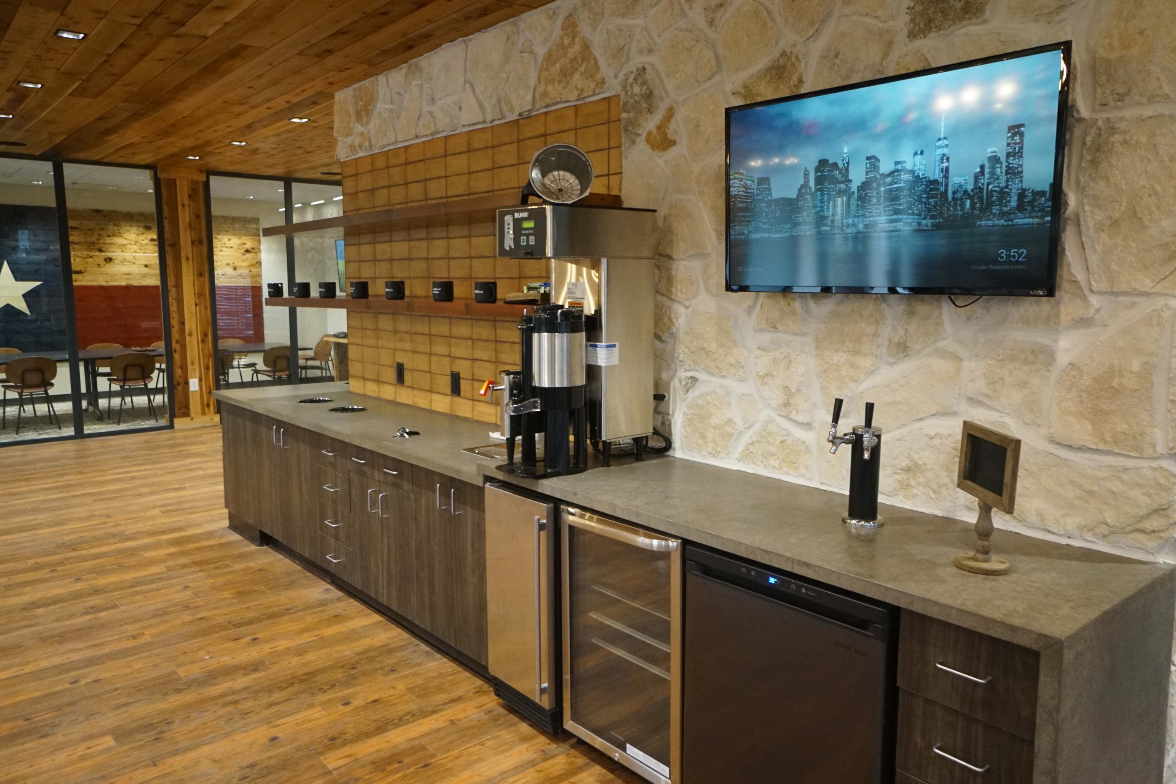 A modern kitchenette with a coffee machine, a refrigerator, and a mounted television, set against a stone wall and wooden flooring.