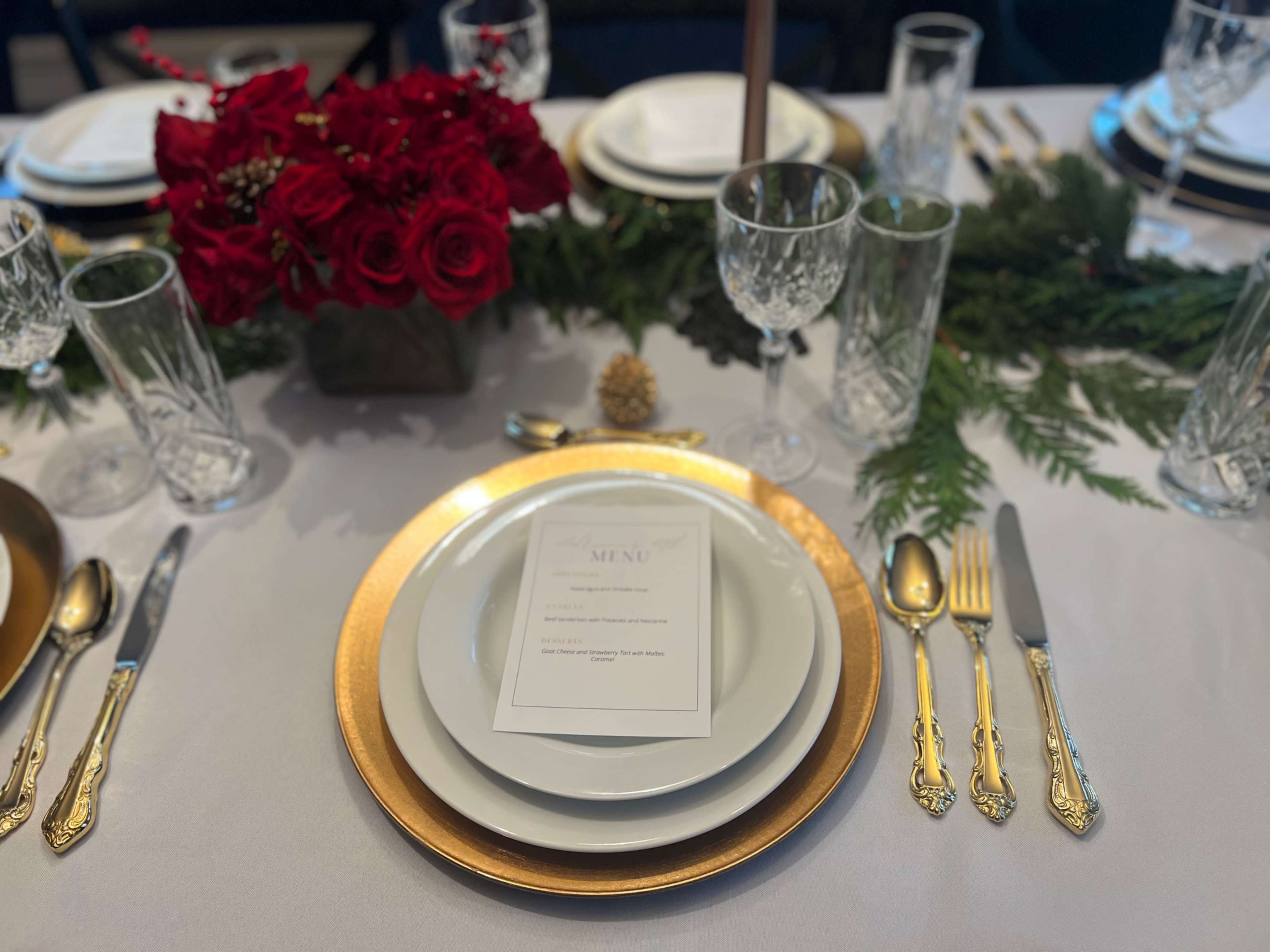 A beautifully arranged dining table features a gold-rimmed plate, a menu card, crystal glassware, silver utensils, and a centerpiece of red roses surrounded by greenery.