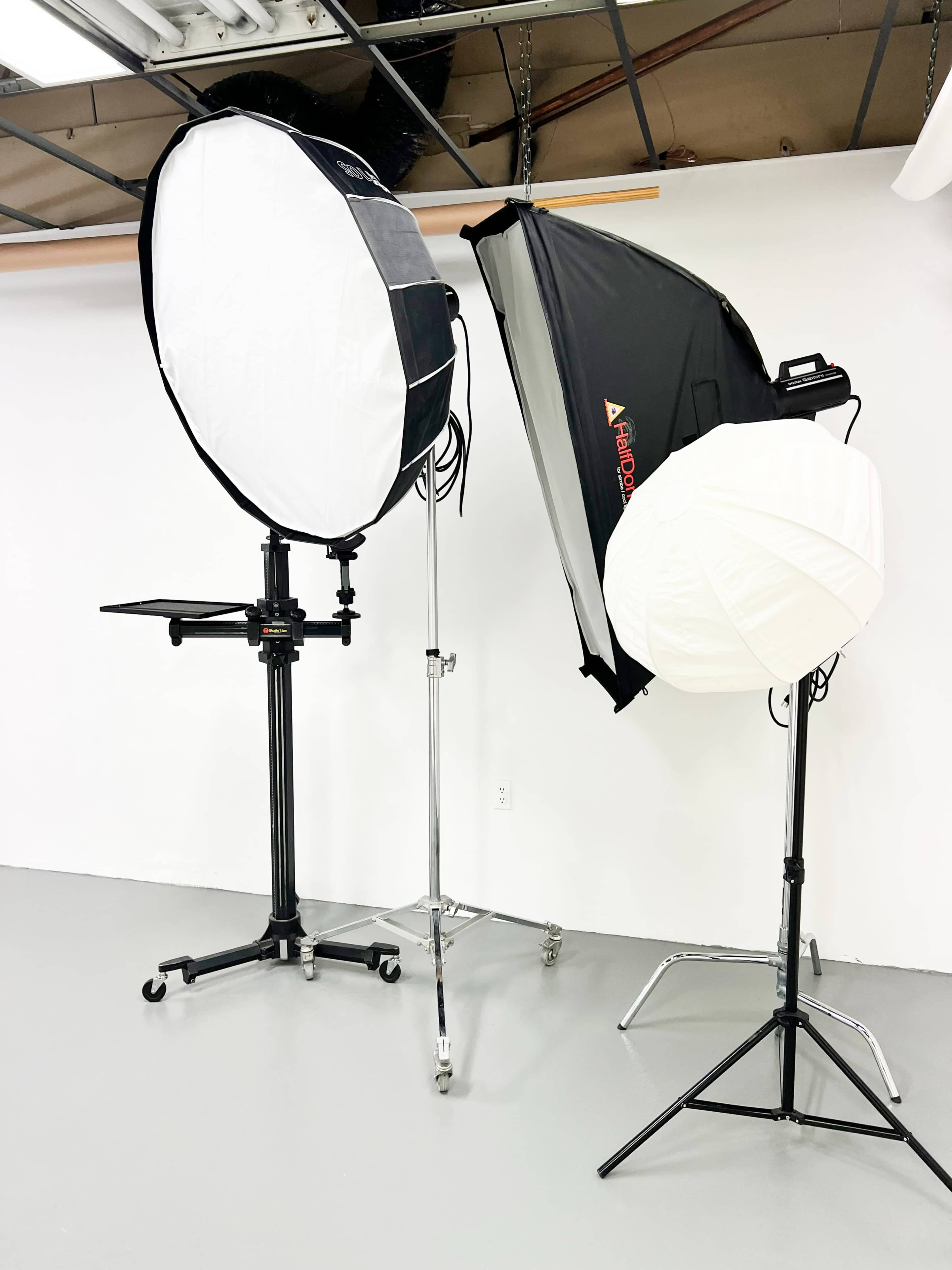 The image shows two photography light setups, including a large round softbox and a smaller white umbrella softbox, both on adjustable stands in a studio environment.