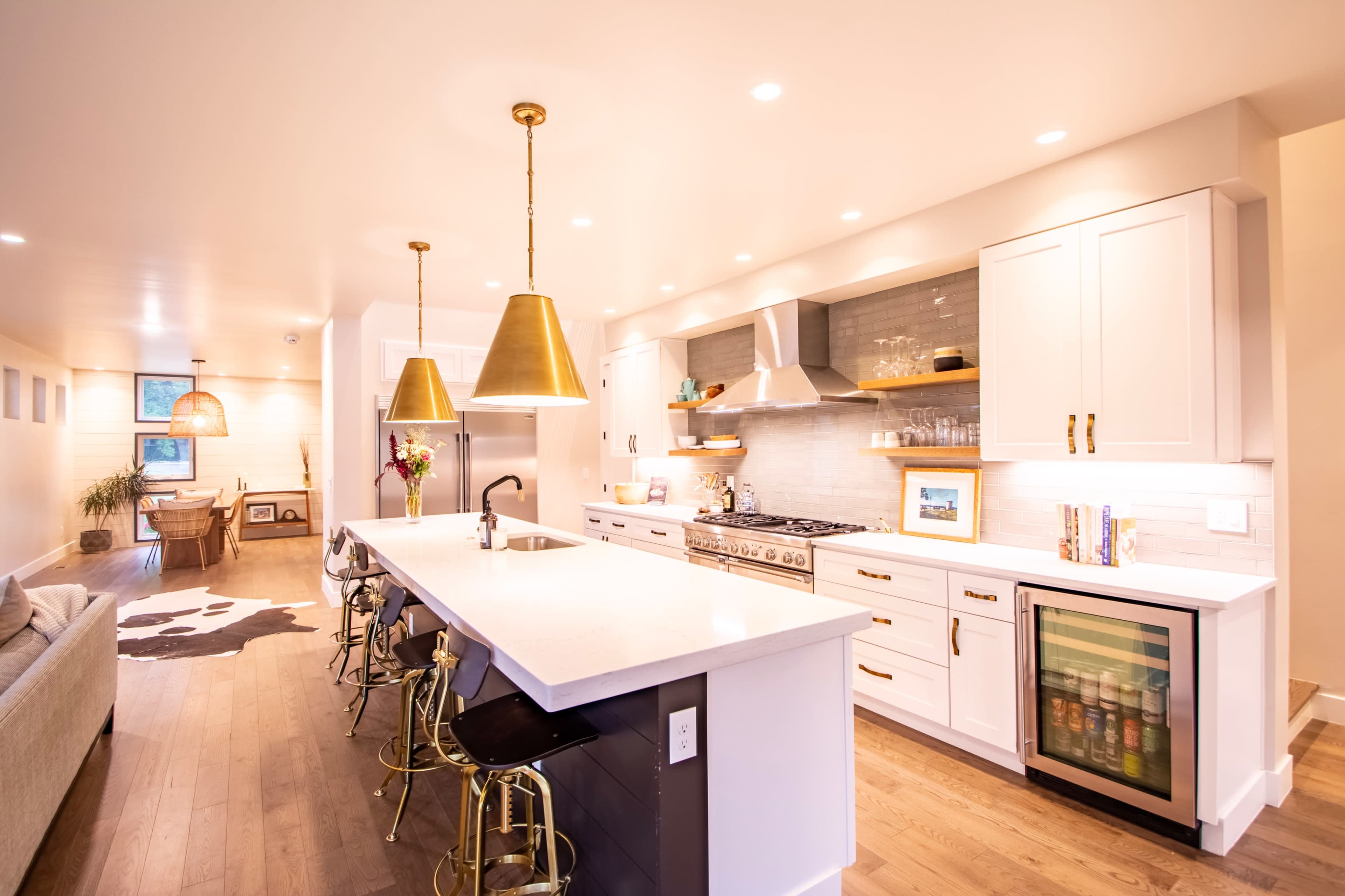 The image shows a modern kitchen featuring a large island with bar stools, gold pendant lights, and stainless steel appliances, all set against light-colored cabinetry and a hardwood floor.