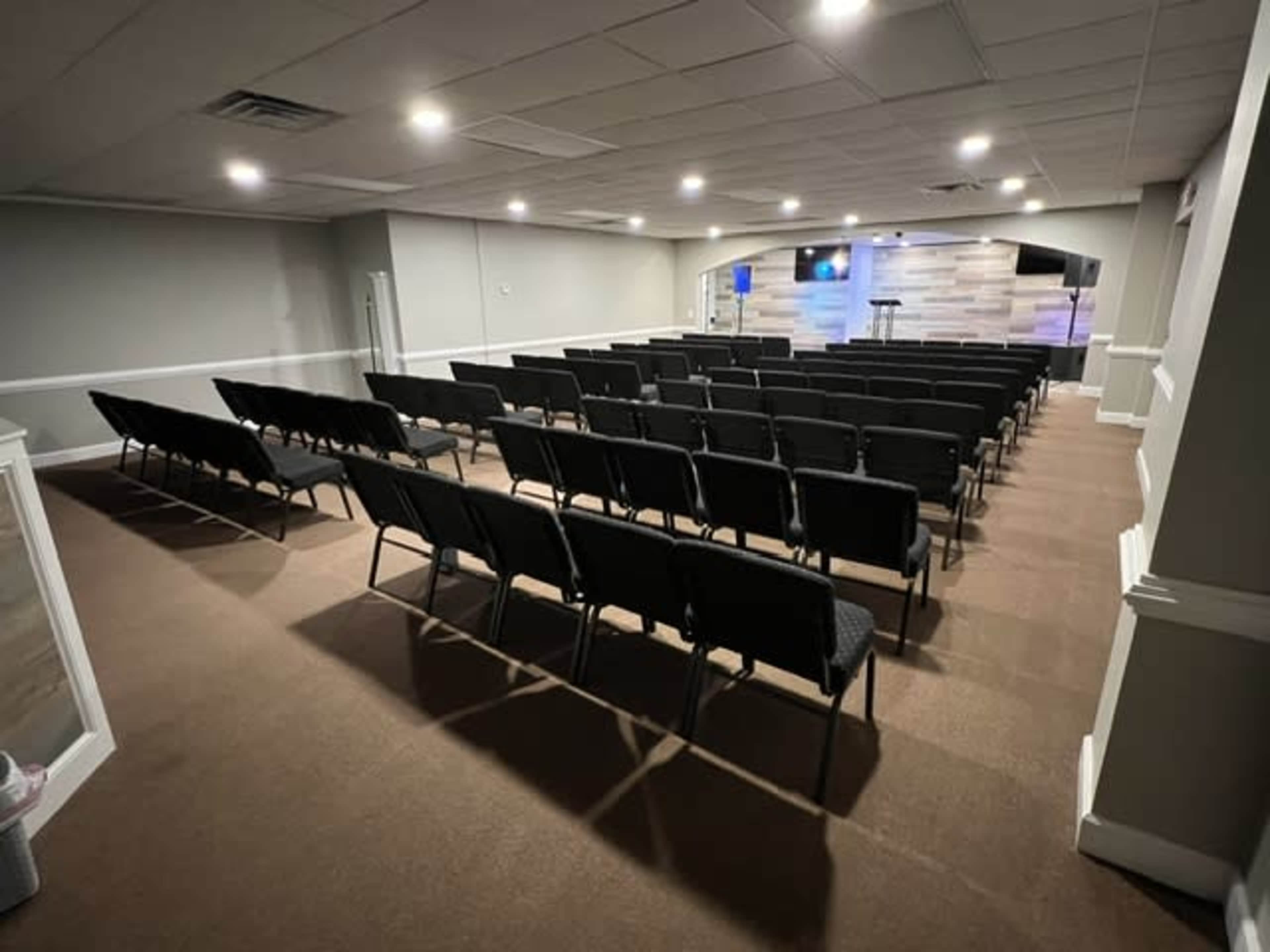 A spacious room with rows of black chairs arranged toward a stage in the front, illuminated by soft lighting.