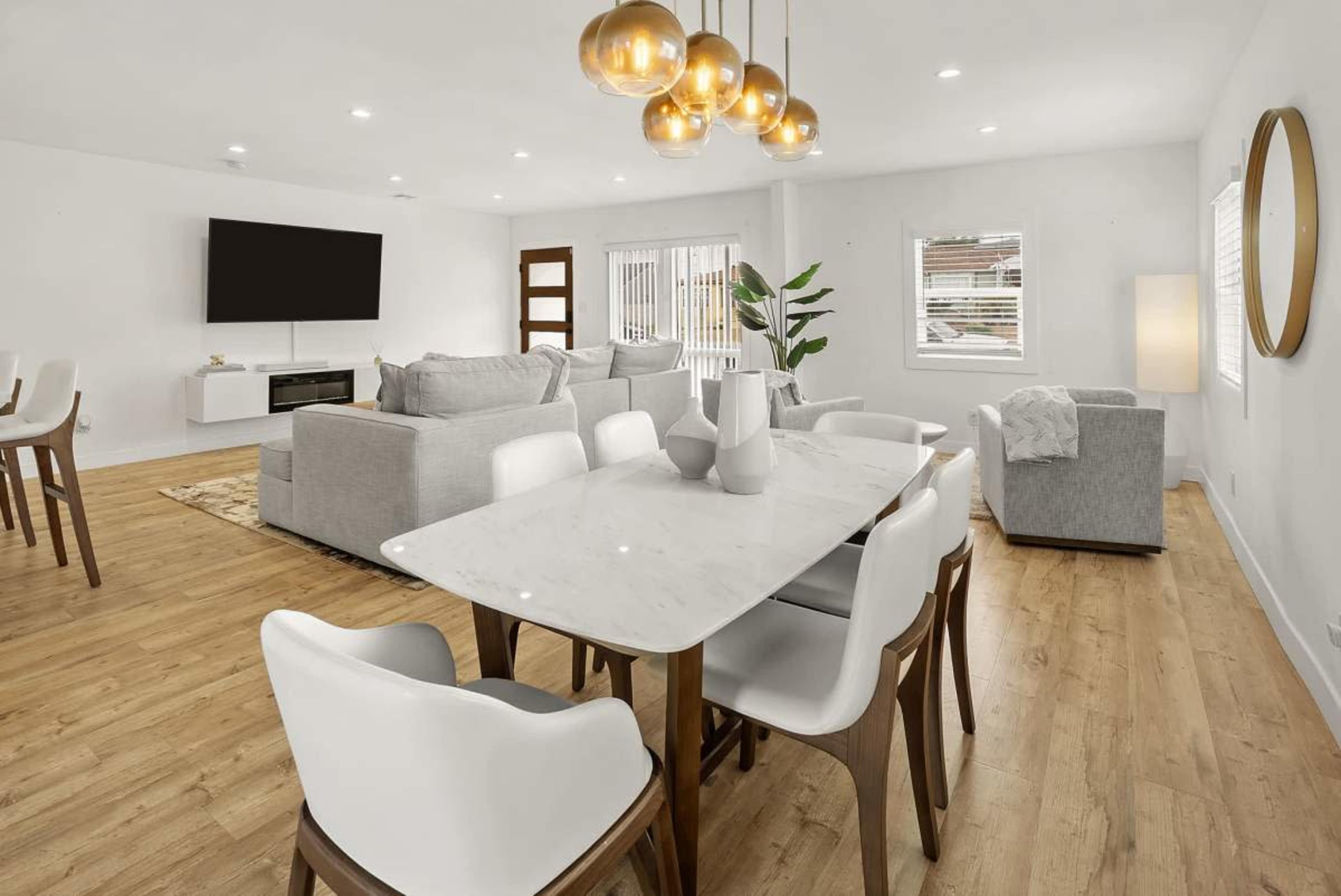 The image shows a modern living and dining area featuring a light-colored sofa, a dining table with white chairs, and pendant lighting above the table.