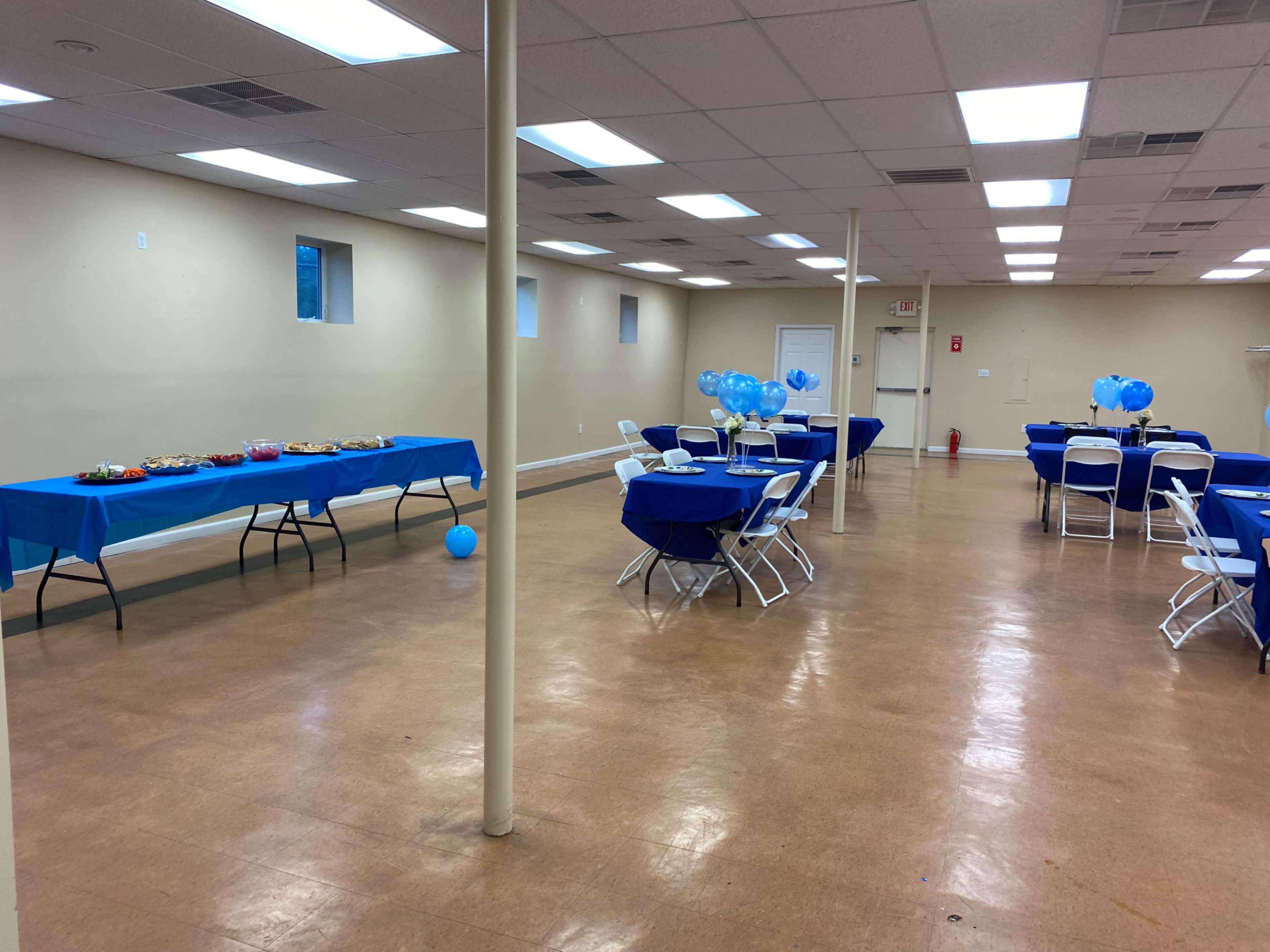 The image shows a spacious room set up for an event, featuring tables with blue tablecloths, decorations, and snacks arranged along one side, while chairs are arranged around smaller tables on the opposite side.