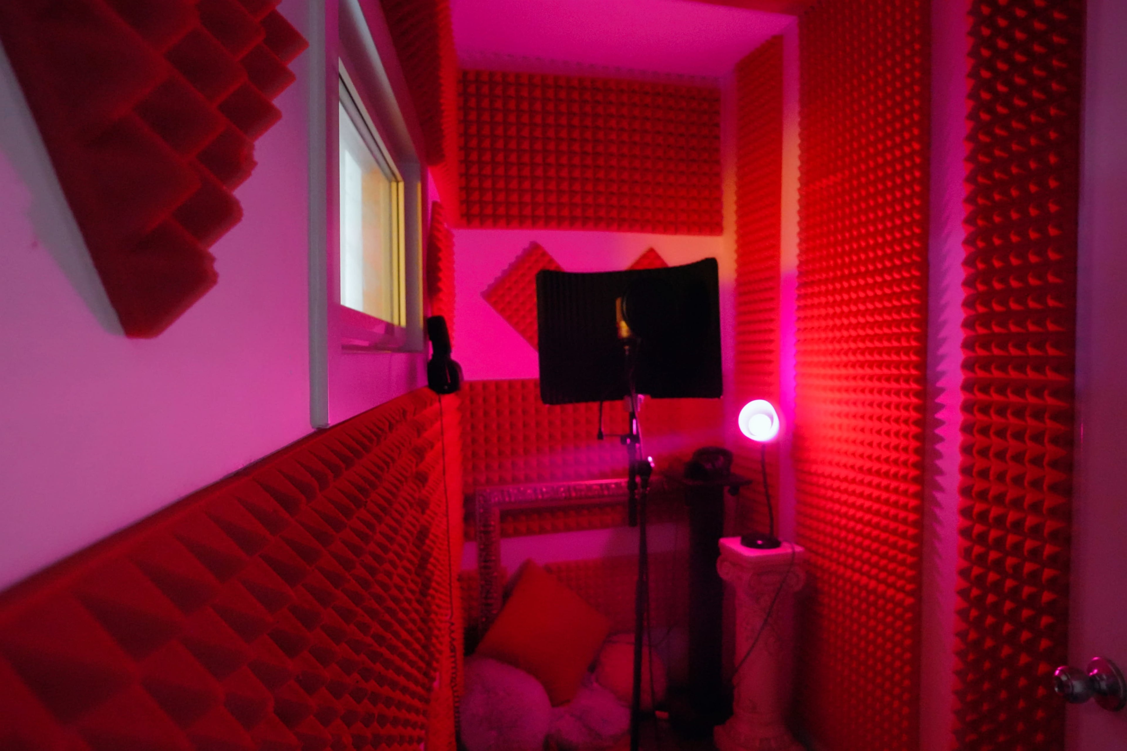 The image shows a soundproof recording booth with red acoustic foam panels on the walls and a microphone setup next to a pink lamp.