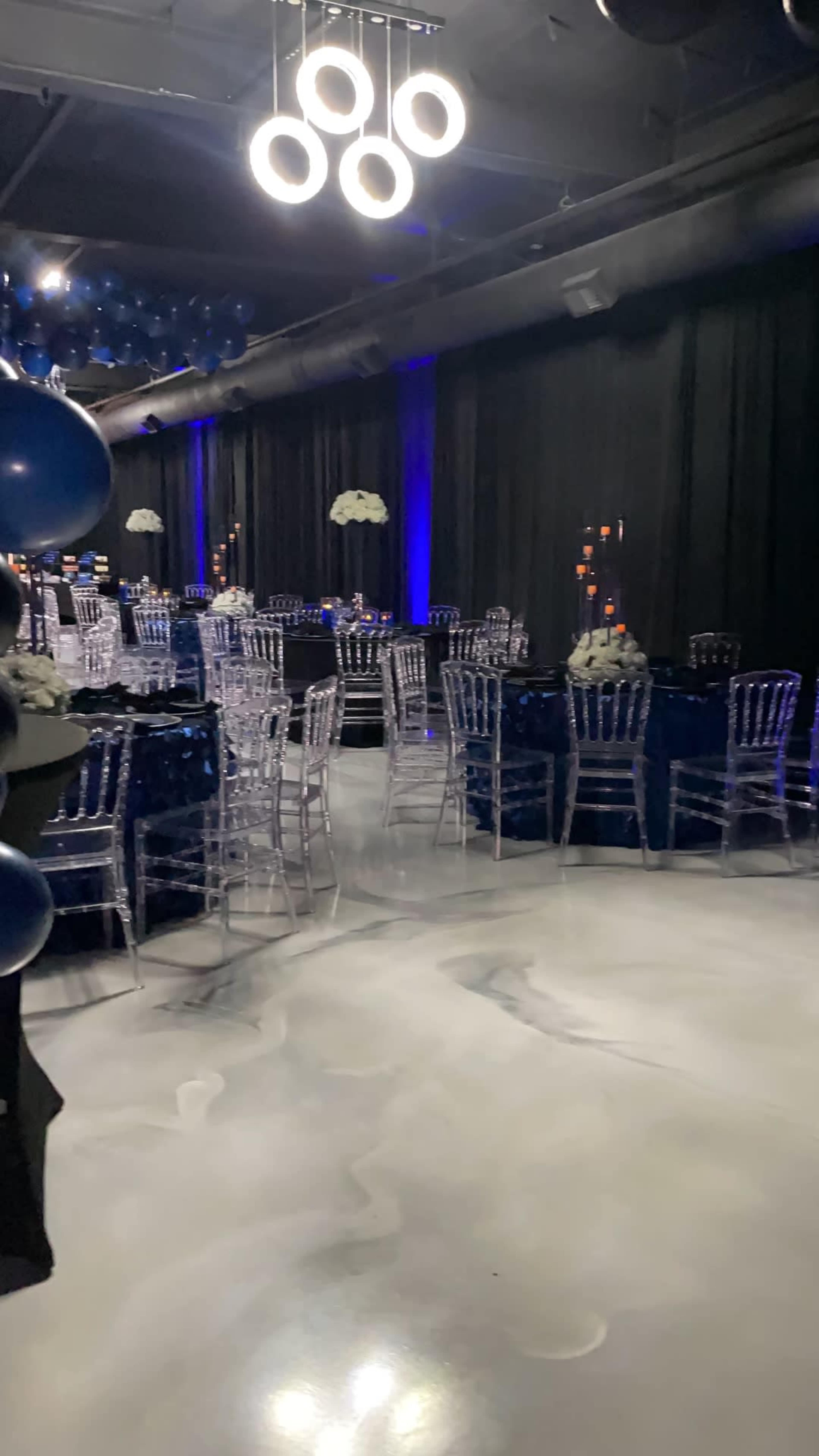 A decorated event space features elegant tables with dark tablecloths, clear chairs, and blue balloons overhead.