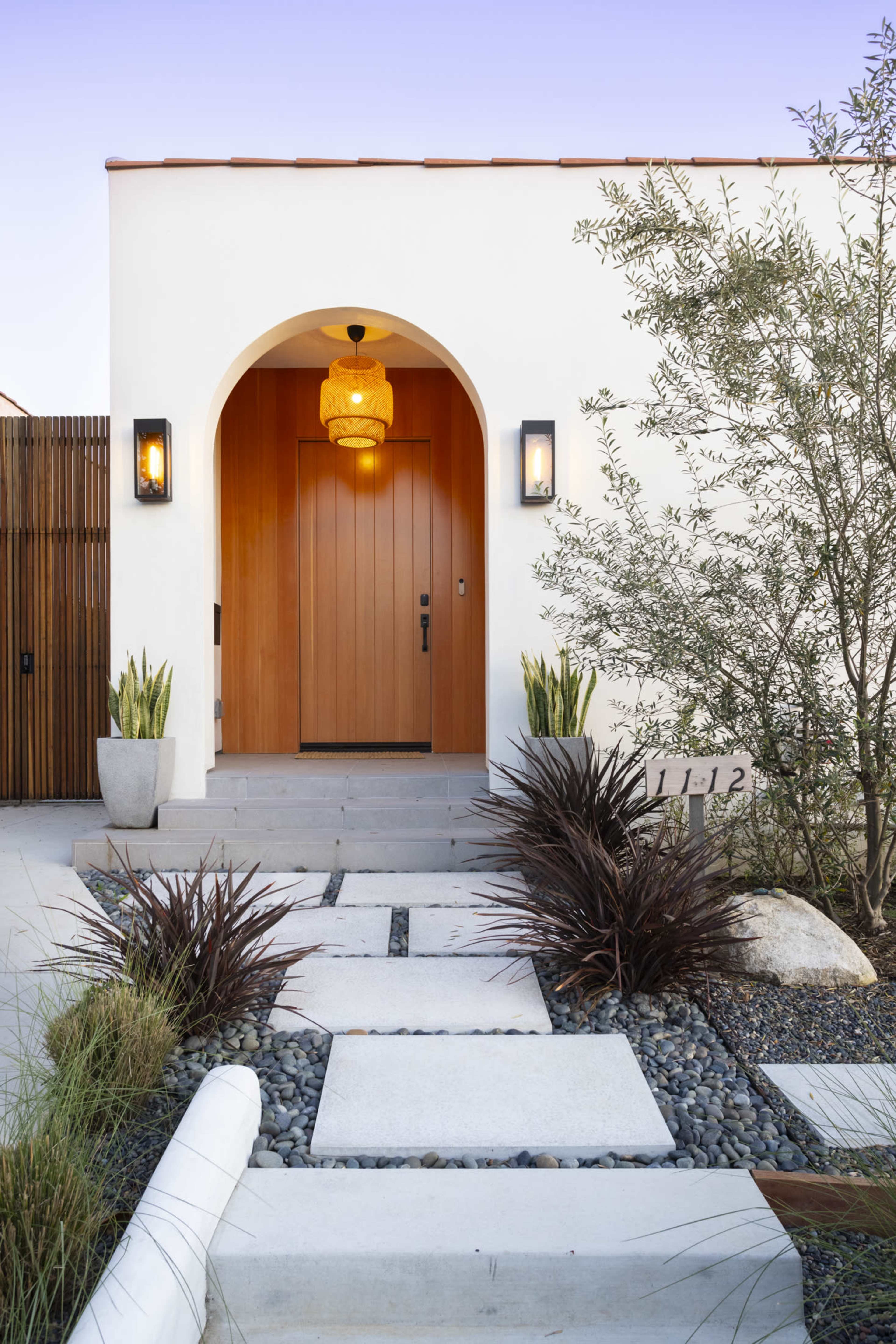 A modern home entrance features a wooden door beneath an arched entryway, flanked by planters and a stone walkway lined with pebbles.
