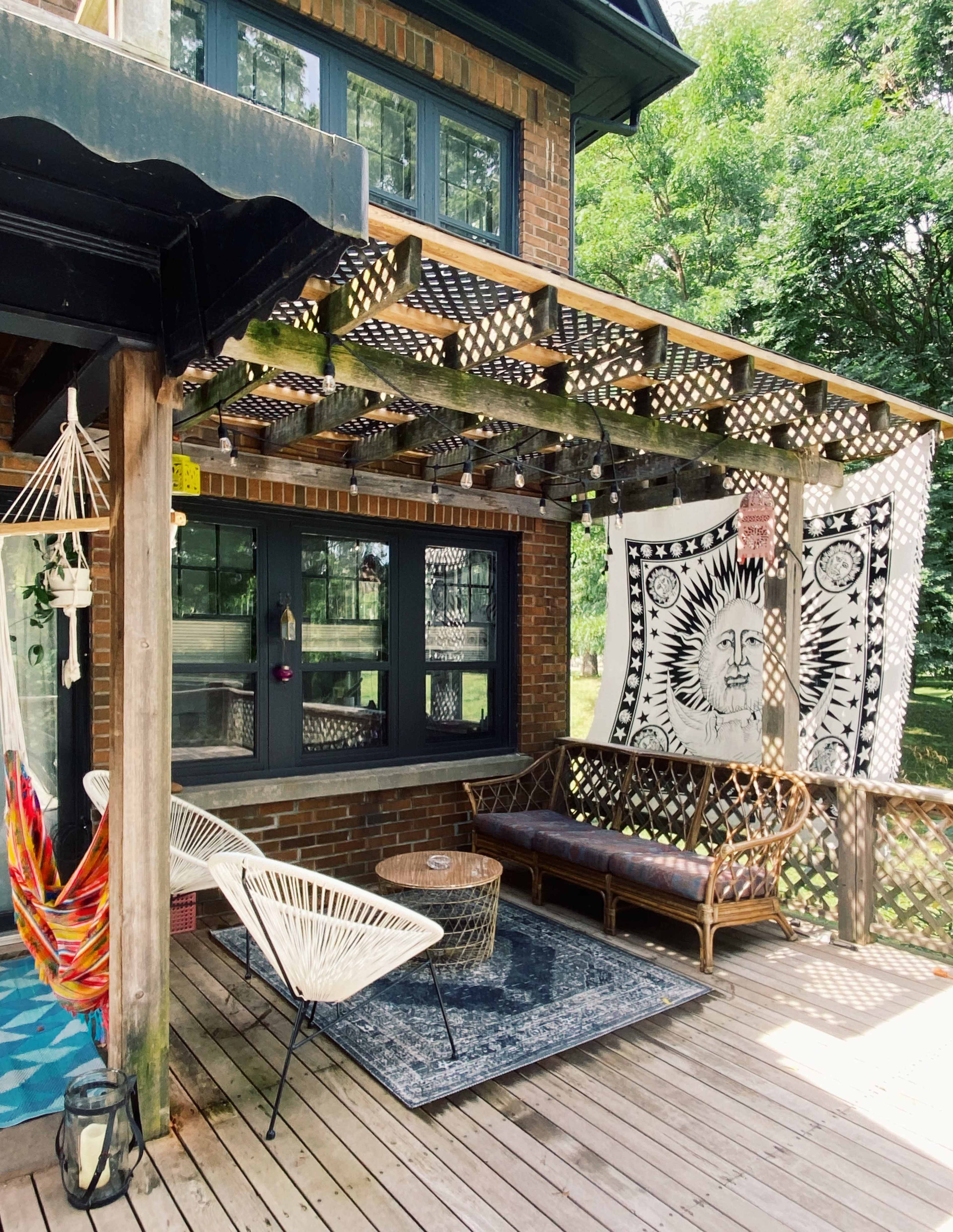 A wooden deck attached to a brick house, featuring a hammock, a rattan sofa, a round table, and a large tapestry hanging on the wall.