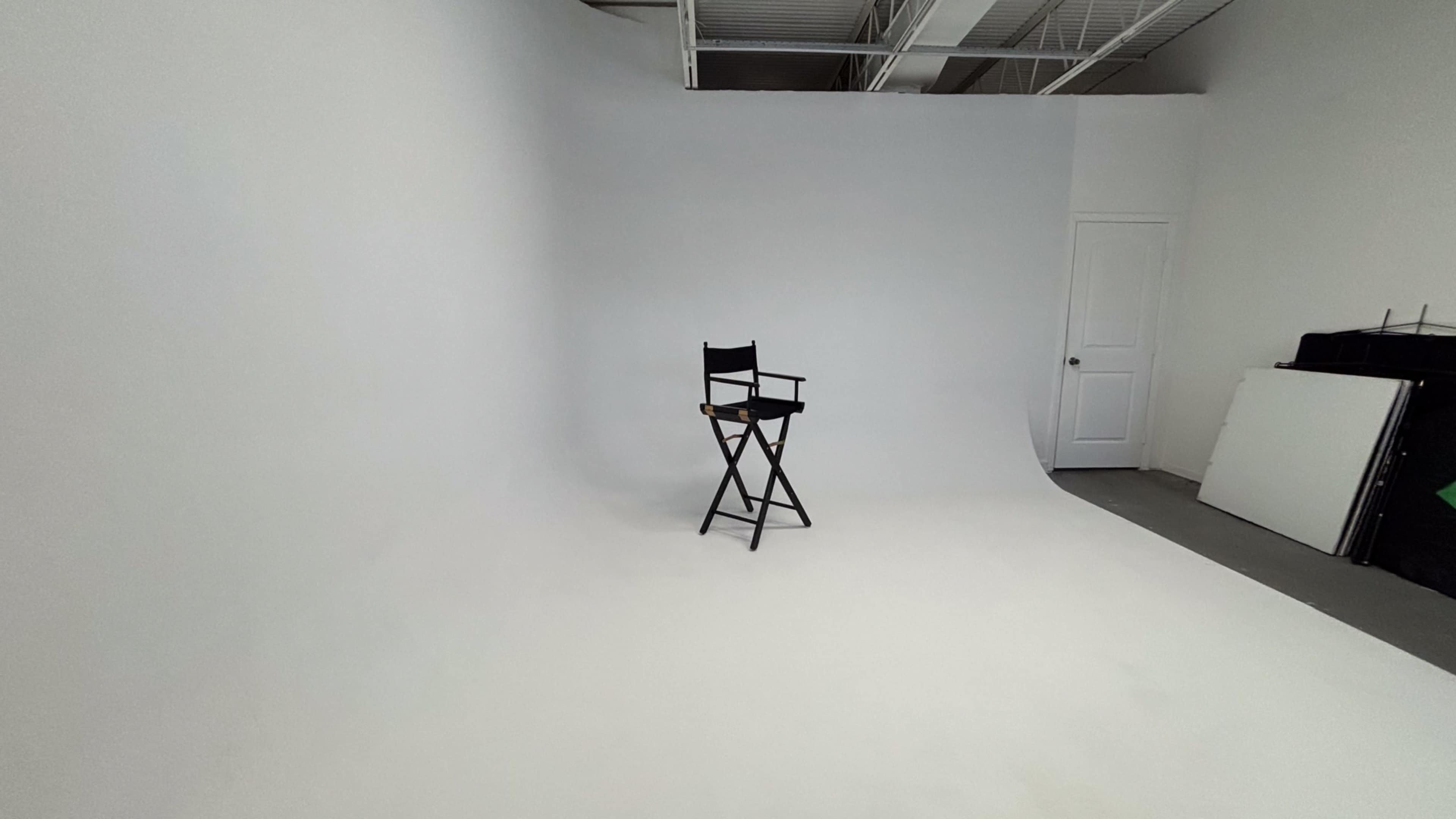 The image shows a minimalist studio space with a blank white backdrop and a single black director's chair positioned in the center.