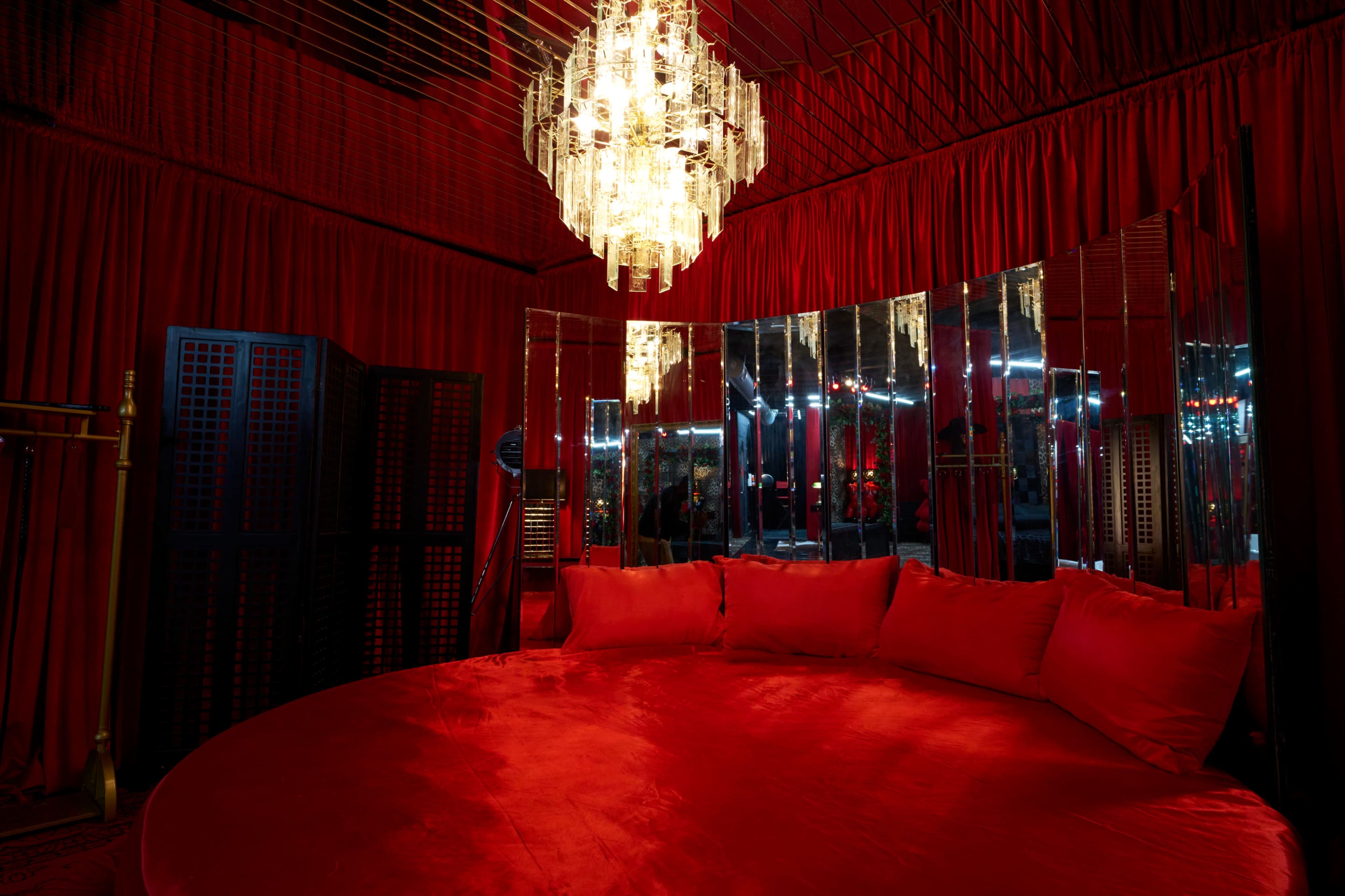 A round red velvet seating area is illuminated by a crystal chandelier, surrounded by mirrored walls and red drapery.