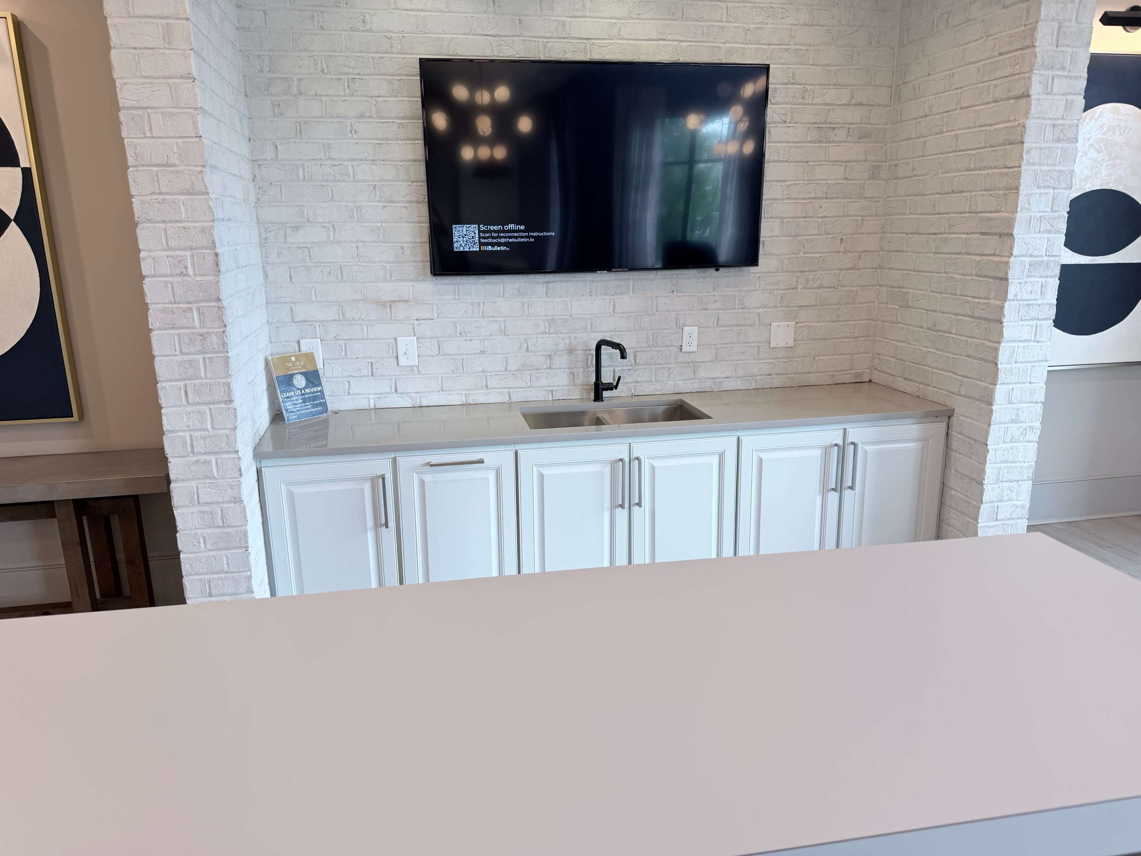 The image shows a modern kitchen area featuring a white brick wall, a built-in television above a sink, and cabinetry below.