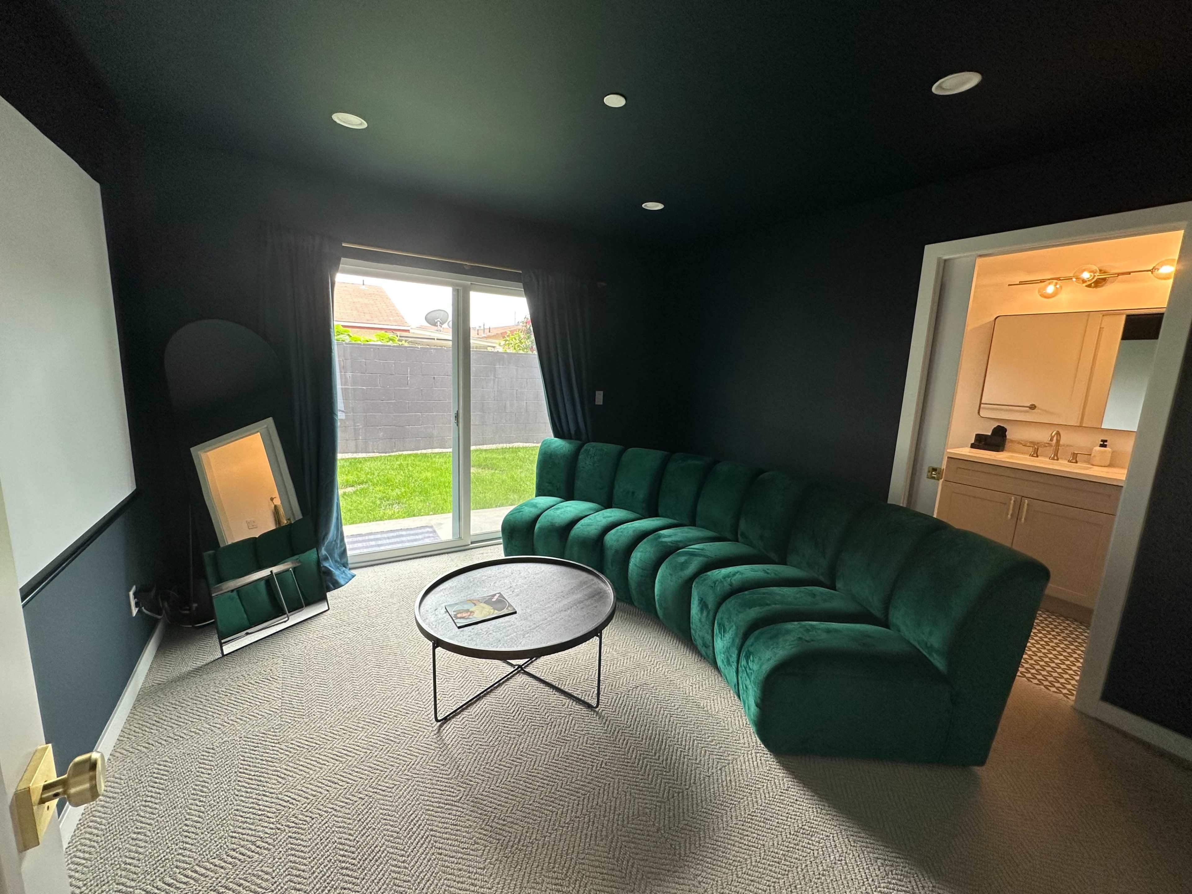 The image shows a darkly painted room with a curved green sofa, a round table, a large mirror, and a door leading to a well-lit area with a sliding glass door opening to a grassy outdoor space.