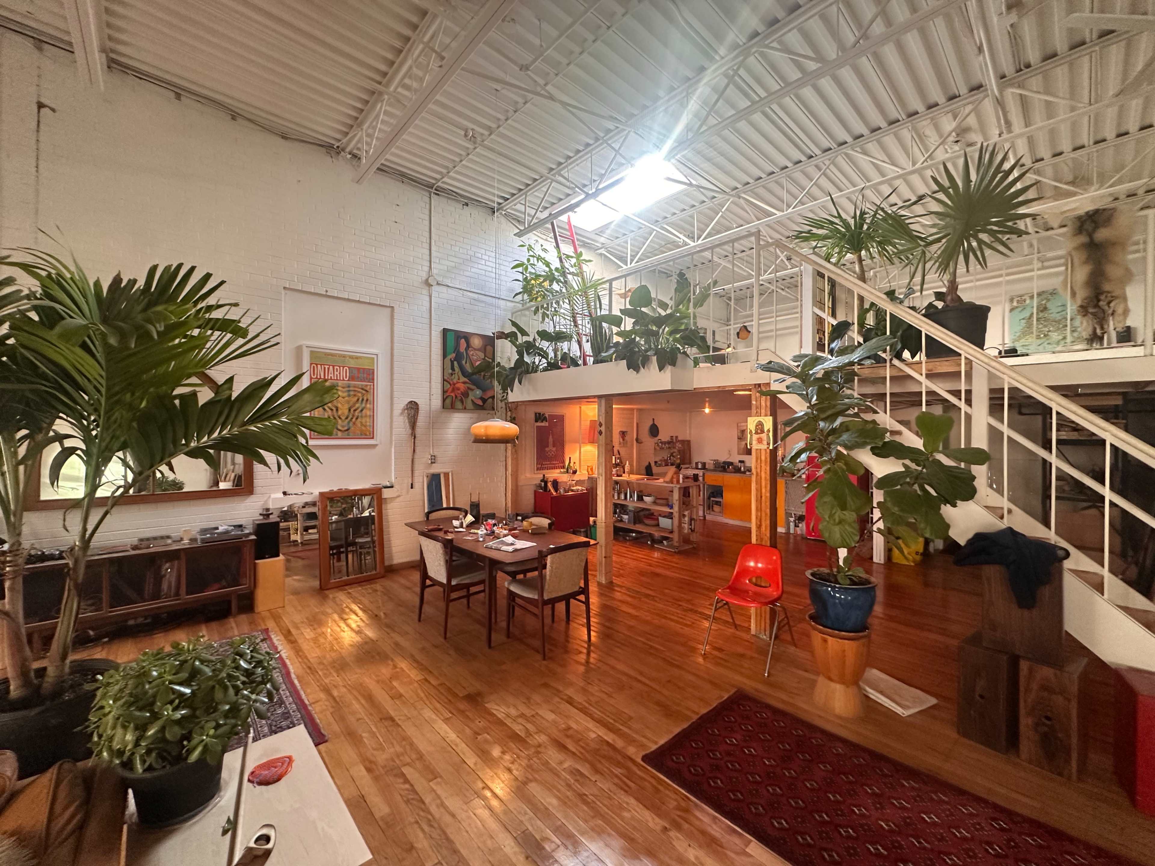 The image shows a spacious, well-lit loft featuring wooden floors, various indoor plants, a dining area with a table and chairs, and a kitchen area in the background.