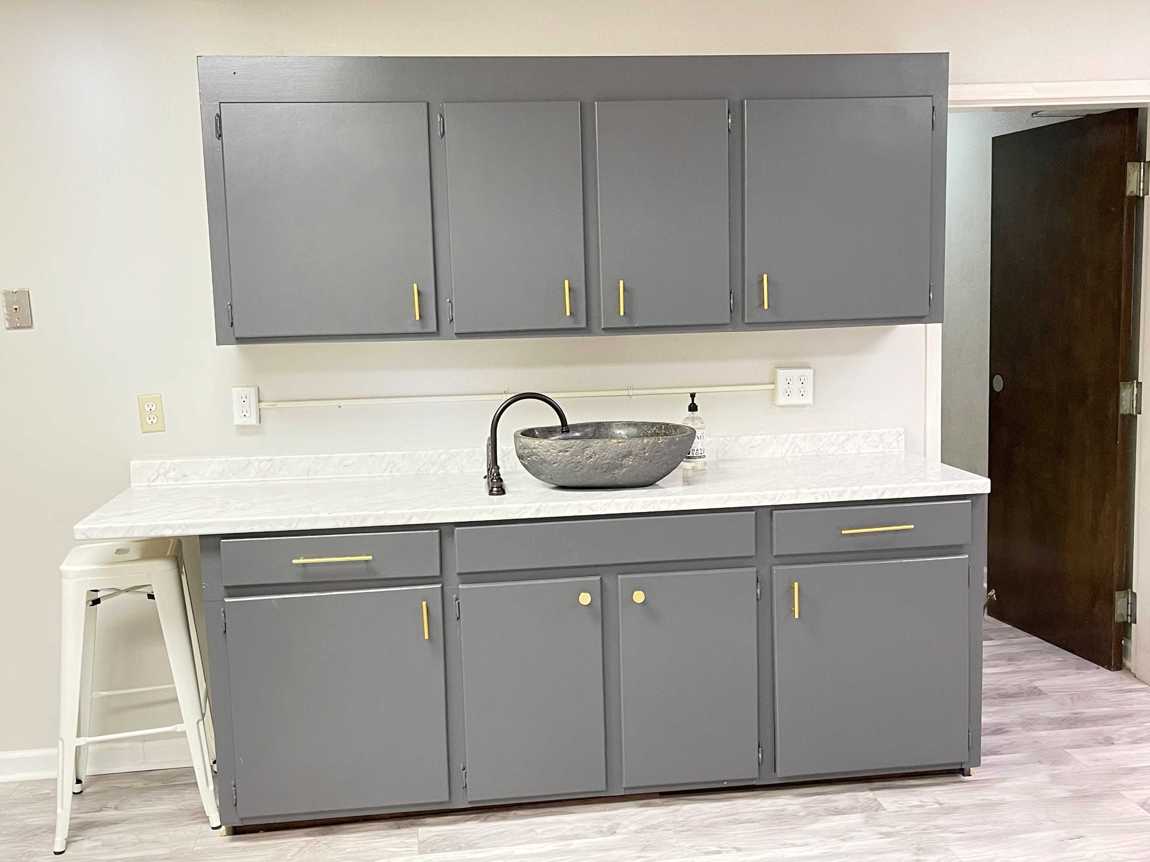 A gray kitchen cabinet set with a marble countertop features a round sink and a nearby white bar stool.