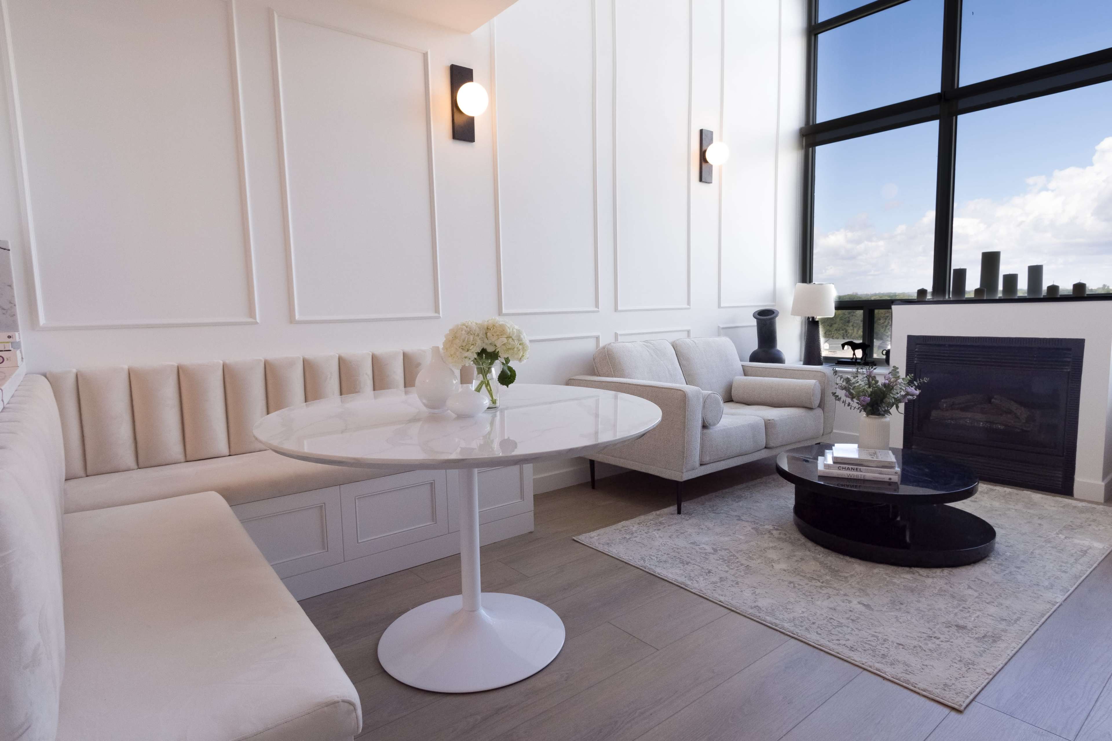 A modern living room with a round table, a plush sofa, and a fireplace, featuring large windows that offer natural light and a view of the sky.