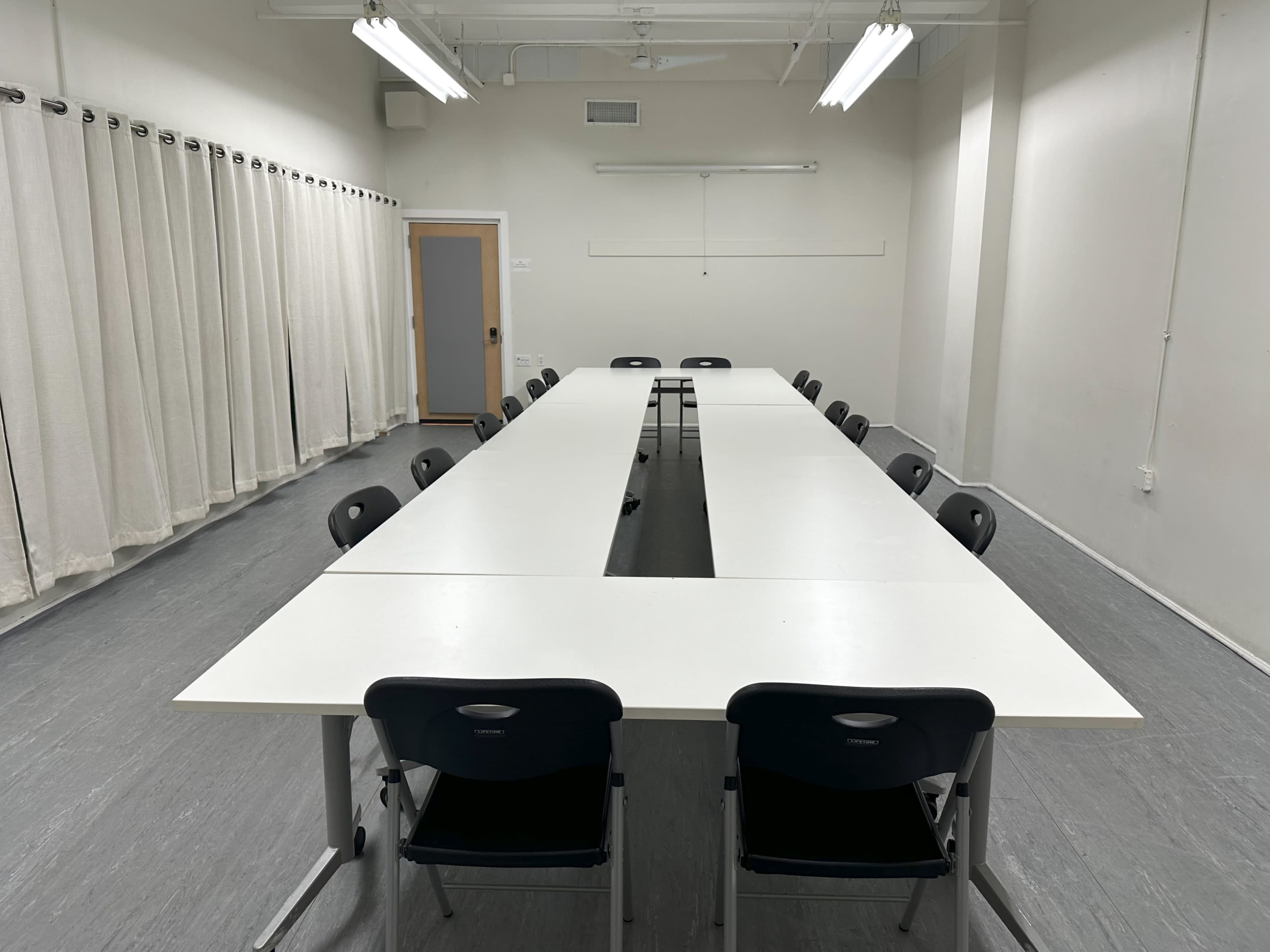 A long, rectangular conference table is set up in a sparsely furnished meeting room surrounded by chairs and curtains.