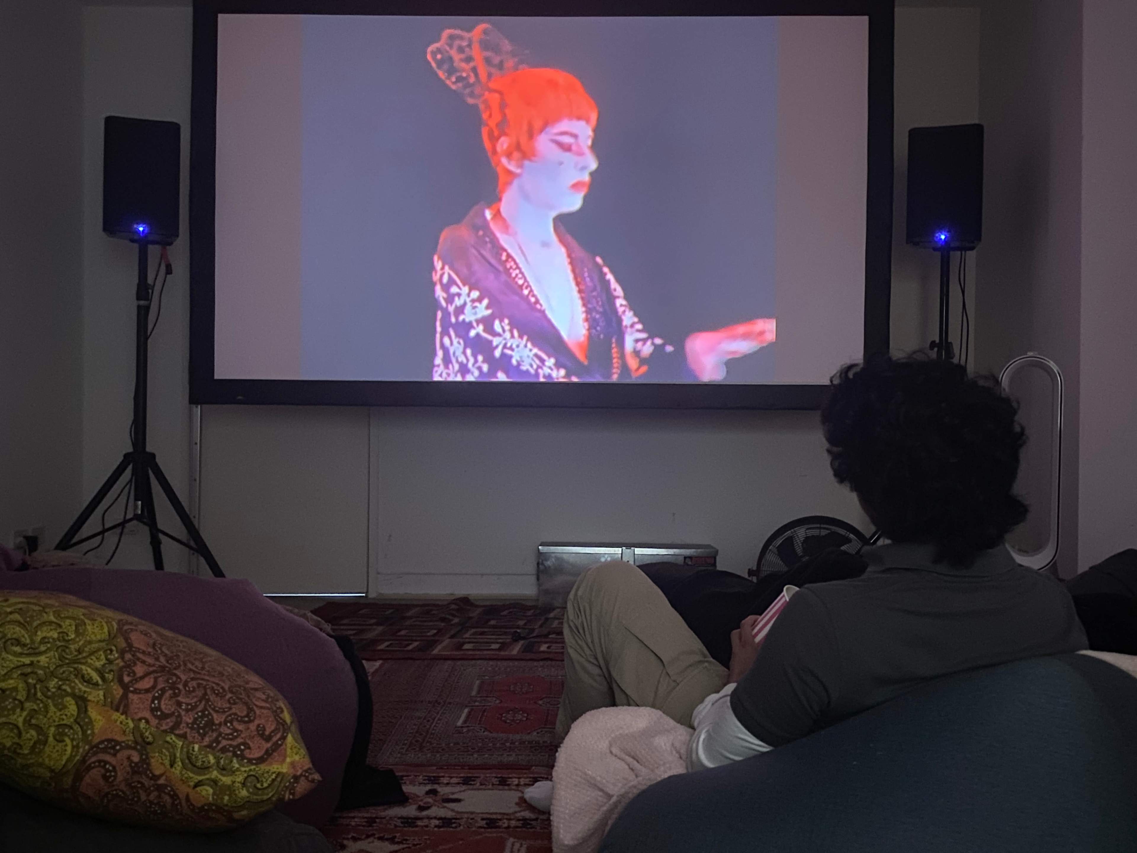 A person sits on a bean bag while watching a colorful performance projected on a screen.
