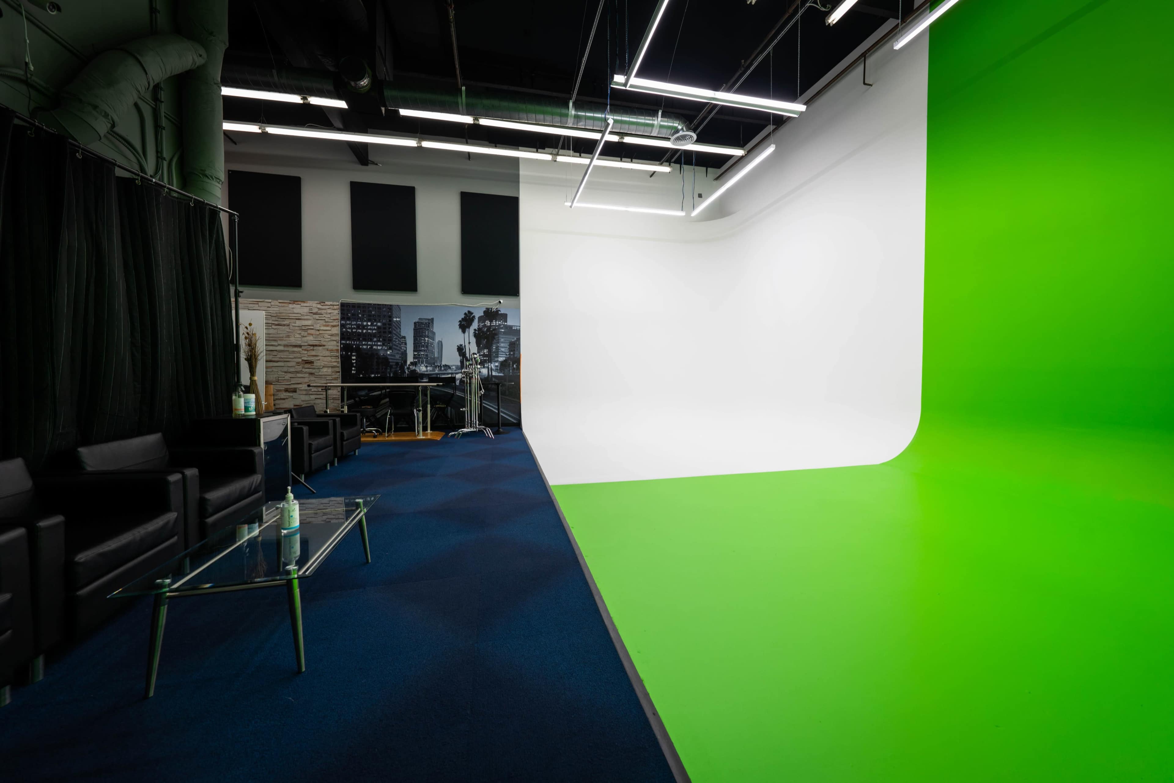 The image shows a photography studio featuring a split backdrop with a white section on one side and a green section on the other, alongside a lounge area with black couches and a glass coffee table.