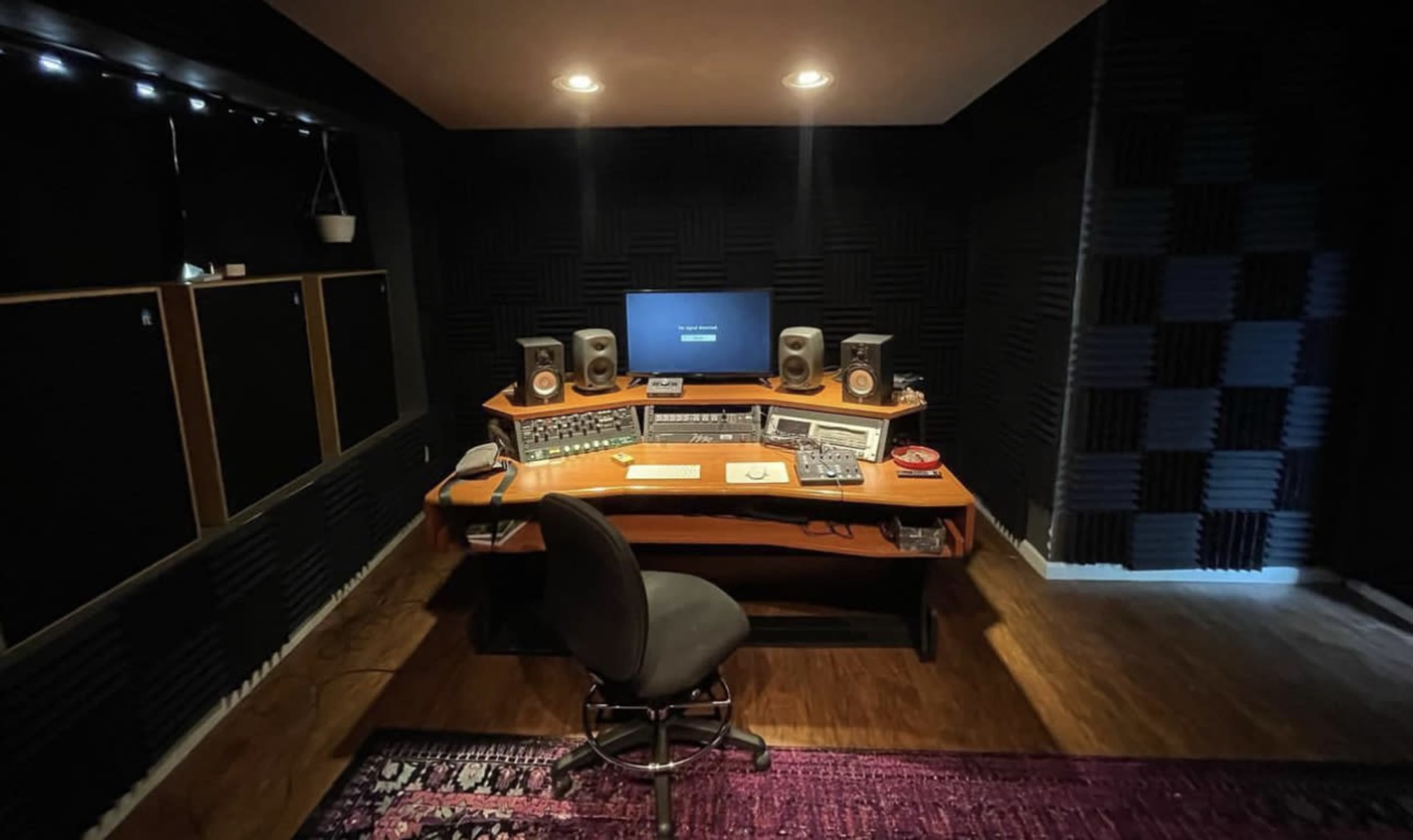 A music recording studio with a wooden desk, computer monitor, speakers, and soundproofing panels on the walls.