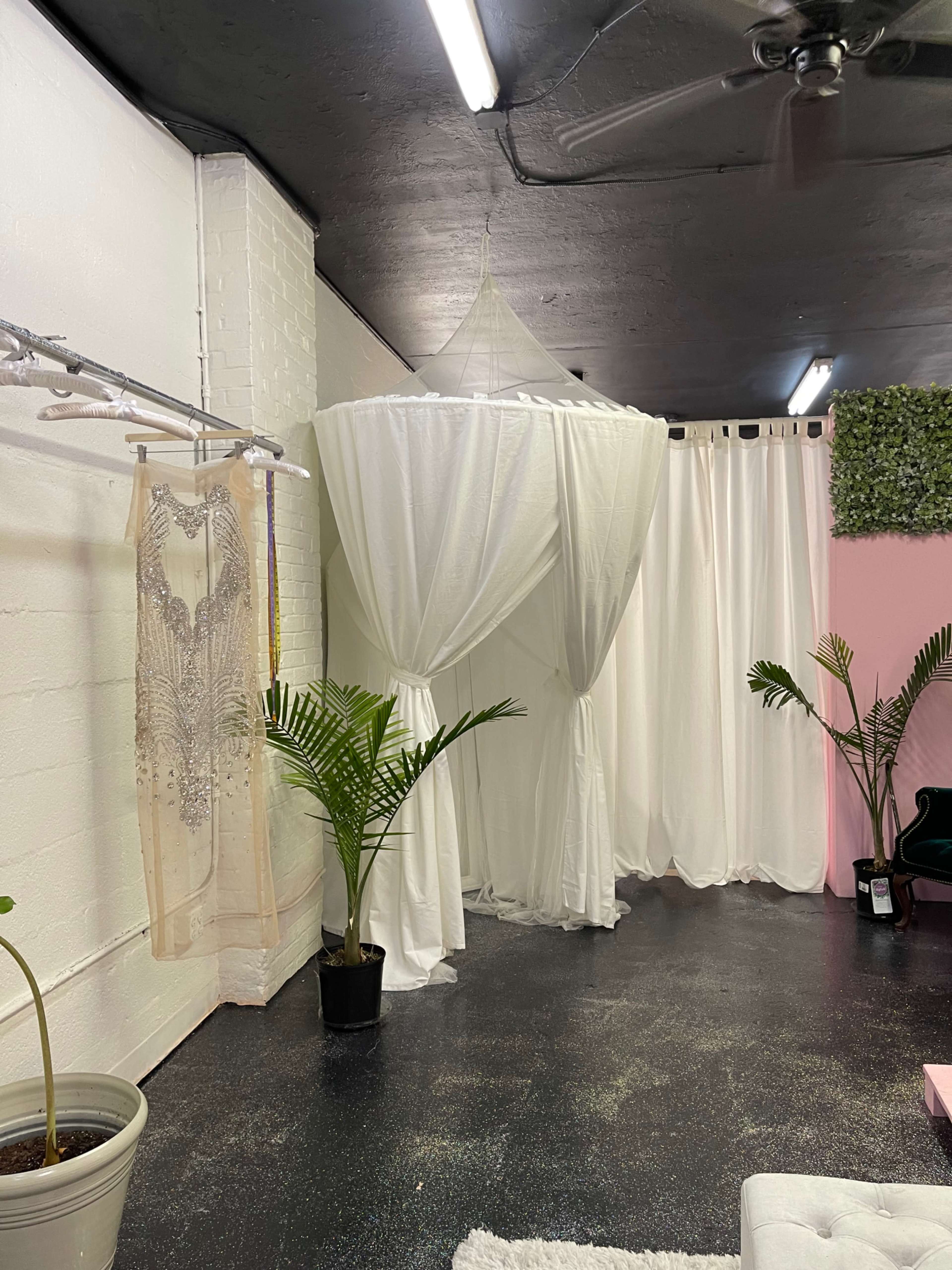 A room with white drapery, a hanging canopy, a potted plant, and a sparkling floor, along with a display of a dress on a rack.