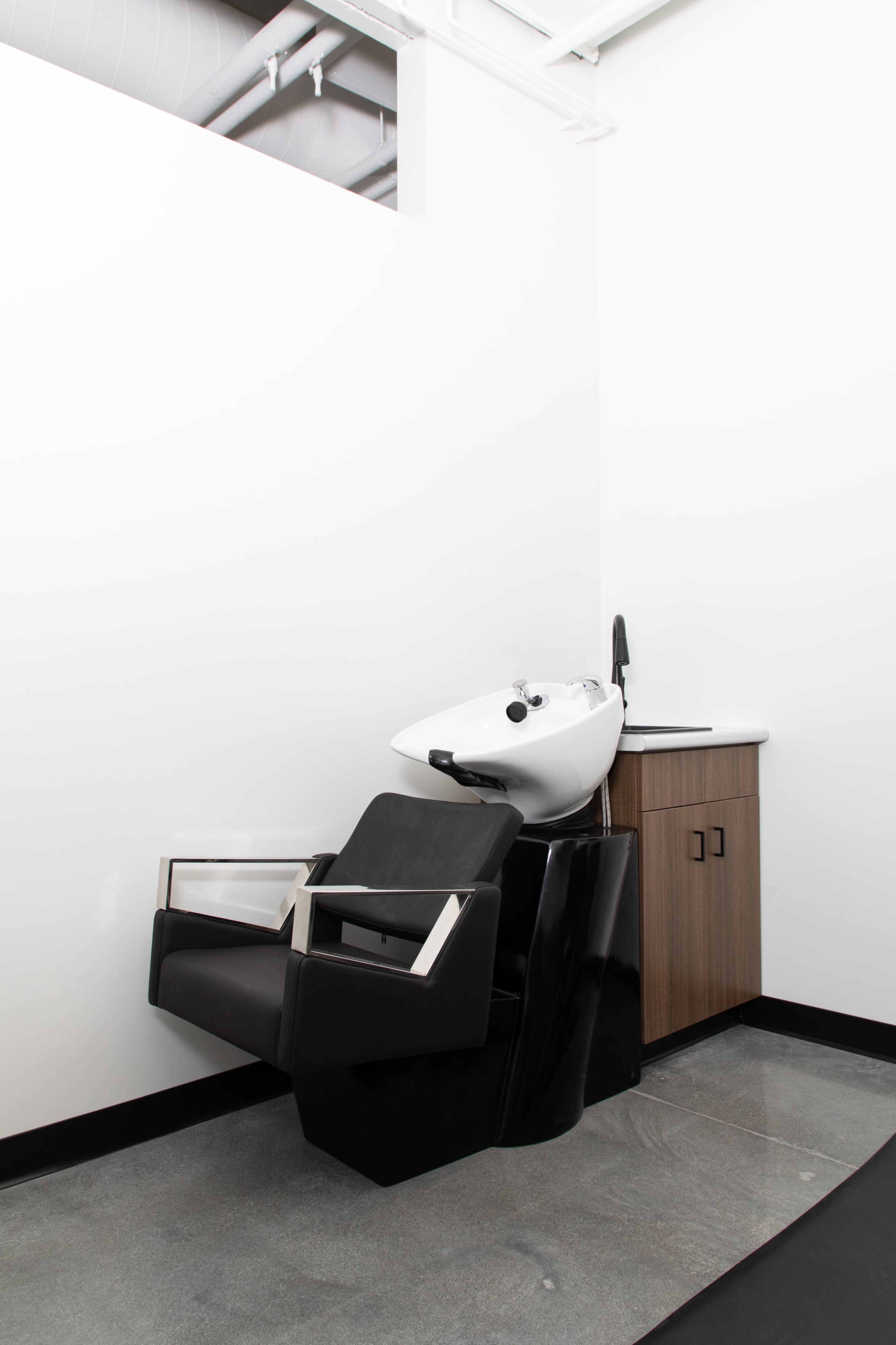 A salon shampoo station with a black reclining chair and a white sink, beside a wooden cabinet, set against a plain white wall.