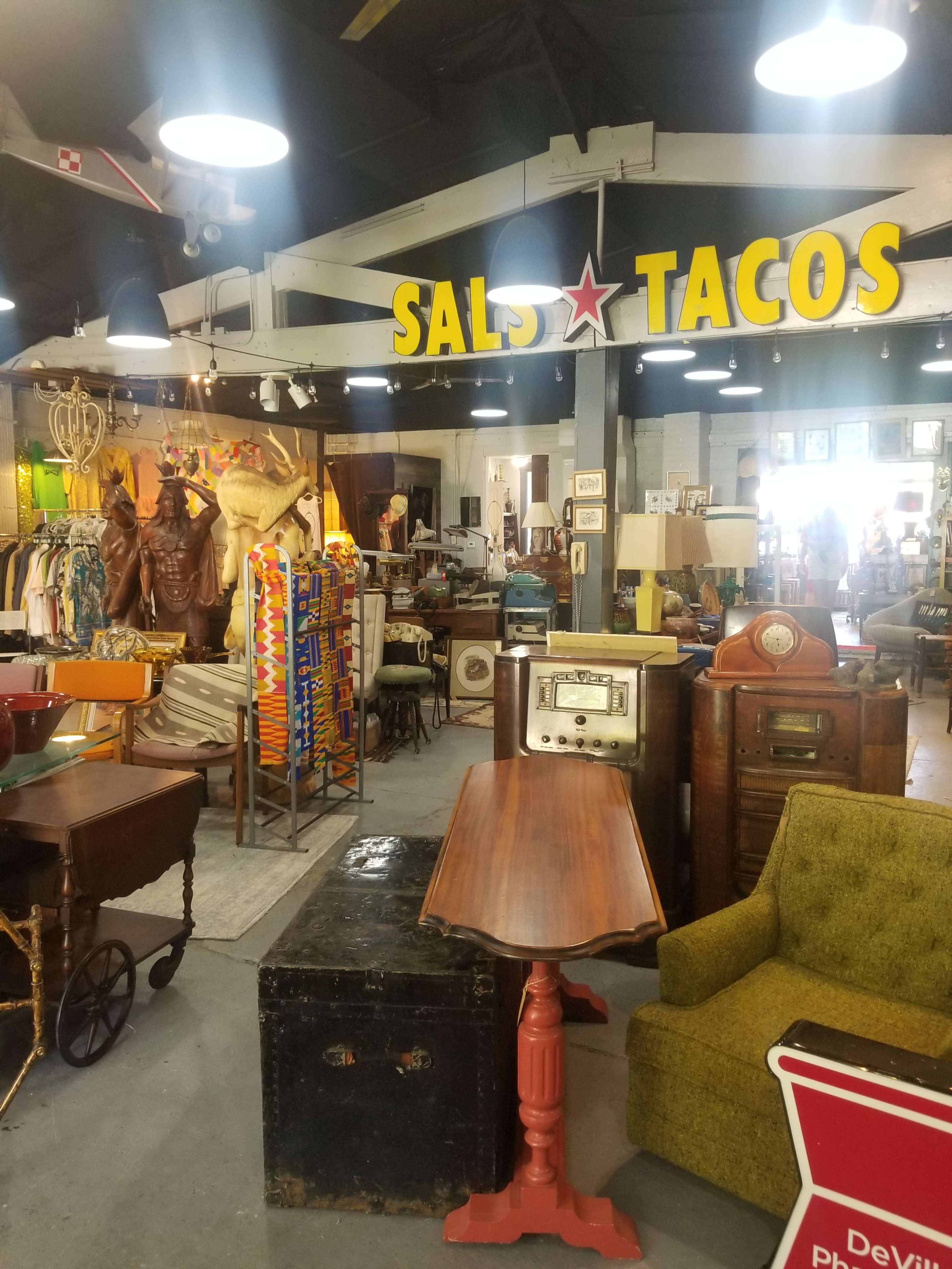 An eclectic antique shop filled with vintage furniture, clothing racks, and various retro items, with a prominent sign that reads "SAL'S TACOS" overhead.