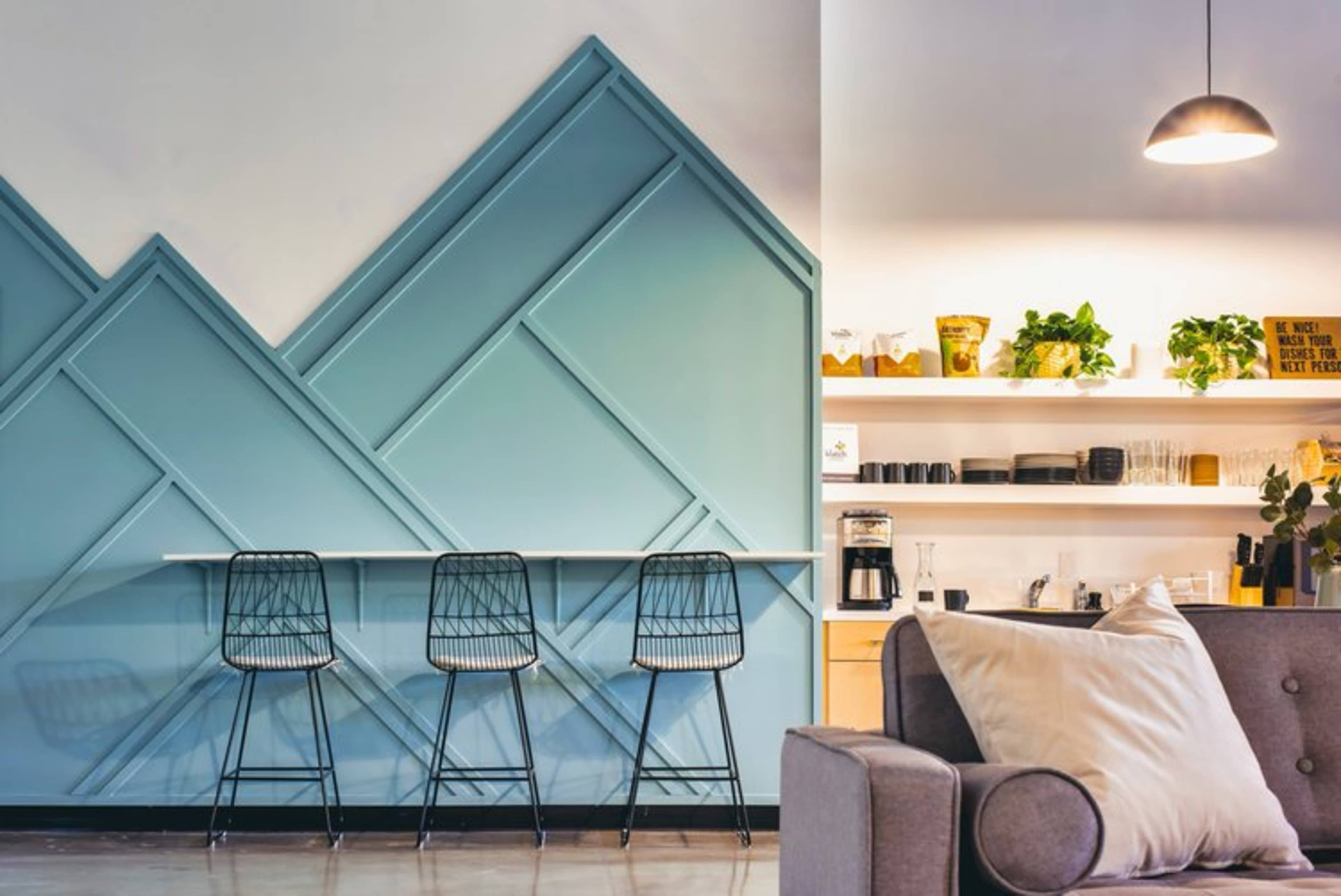 A modern cafe interior with a long bar table against a blue geometric wall, complemented by black wire stools and a shelf displaying plants and kitchenware.