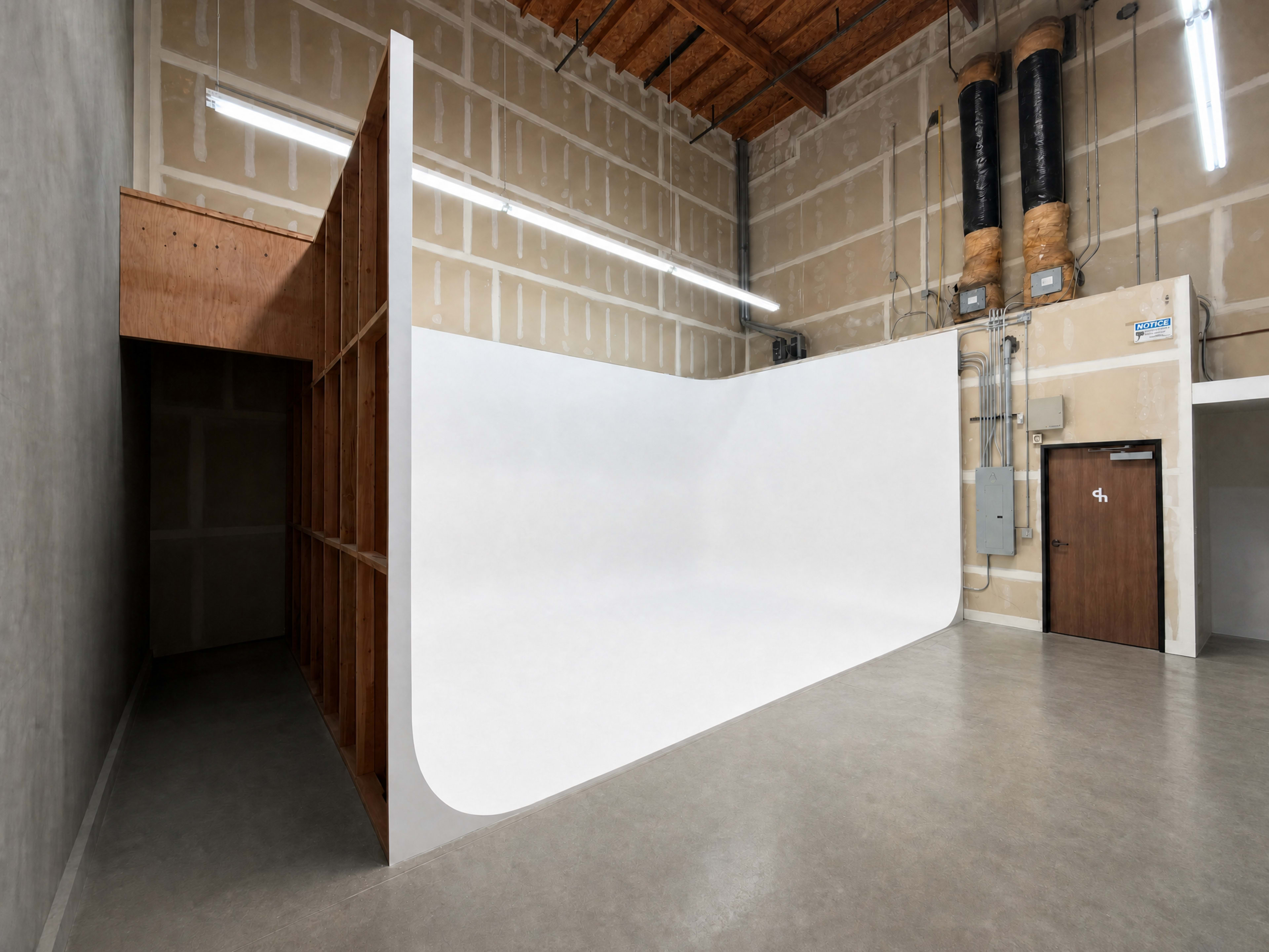 The image shows a spacious indoor area featuring a curved white backdrop mounted against a wooden frame, with exposed walls and ceiling elements in the background.