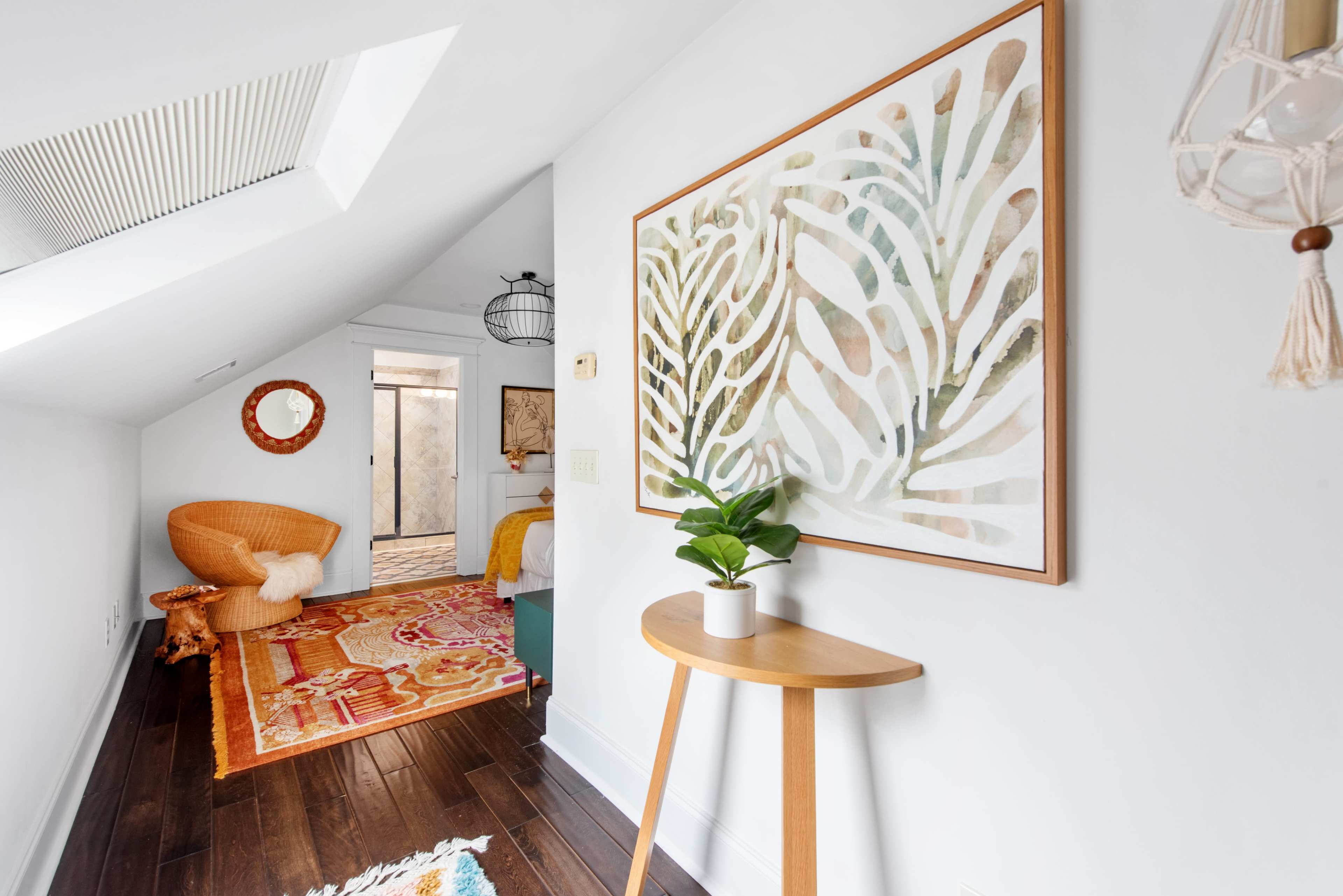 The image shows a bright, small hallway with a decorative patterned rug, a piece of wall art featuring plant motifs, and a small wooden table with a potted plant.