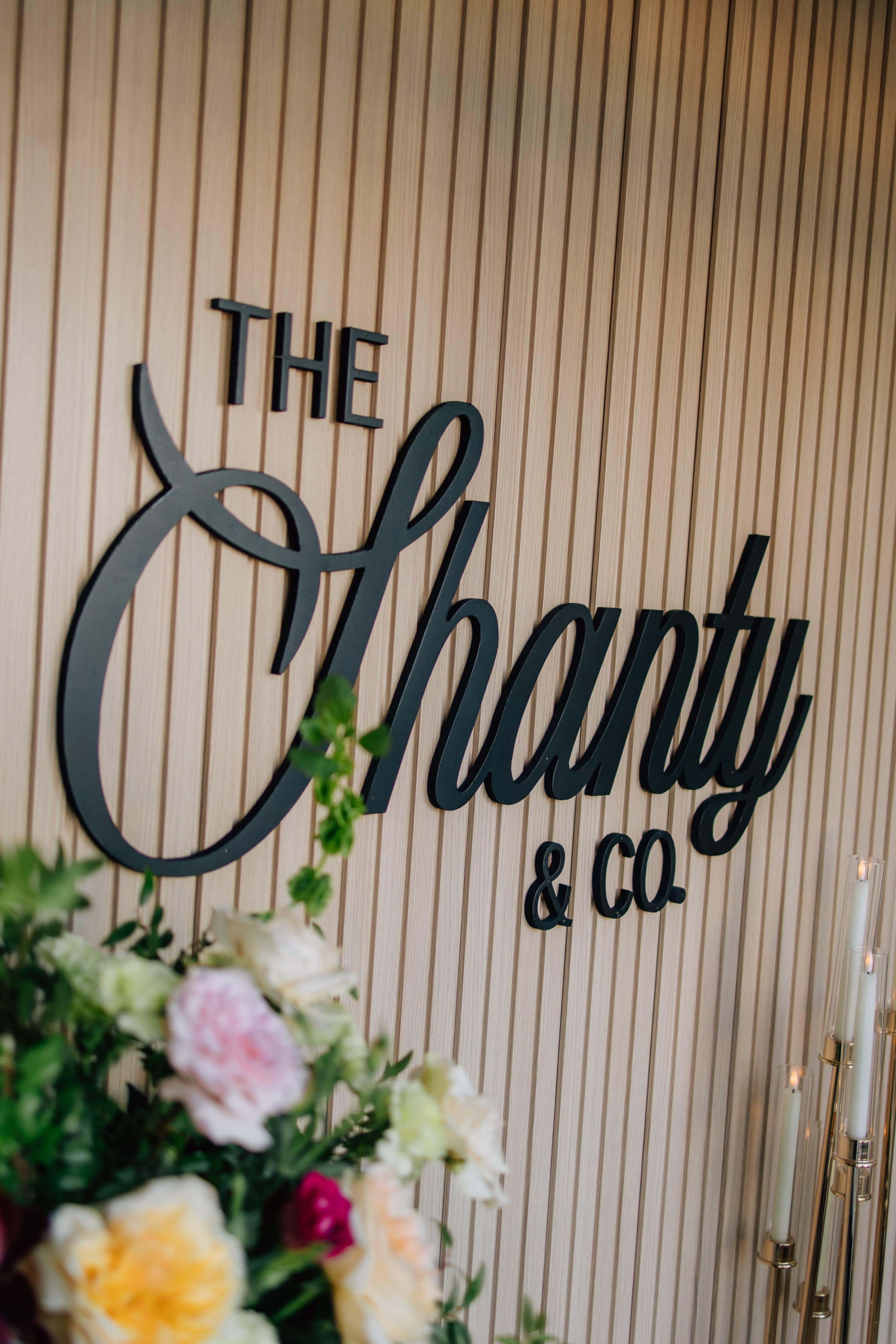 The image features a decorative wall displaying the name "The Shanty & Co" in elegant lettering, accompanied by a arrangement of colorful flowers in the foreground.
