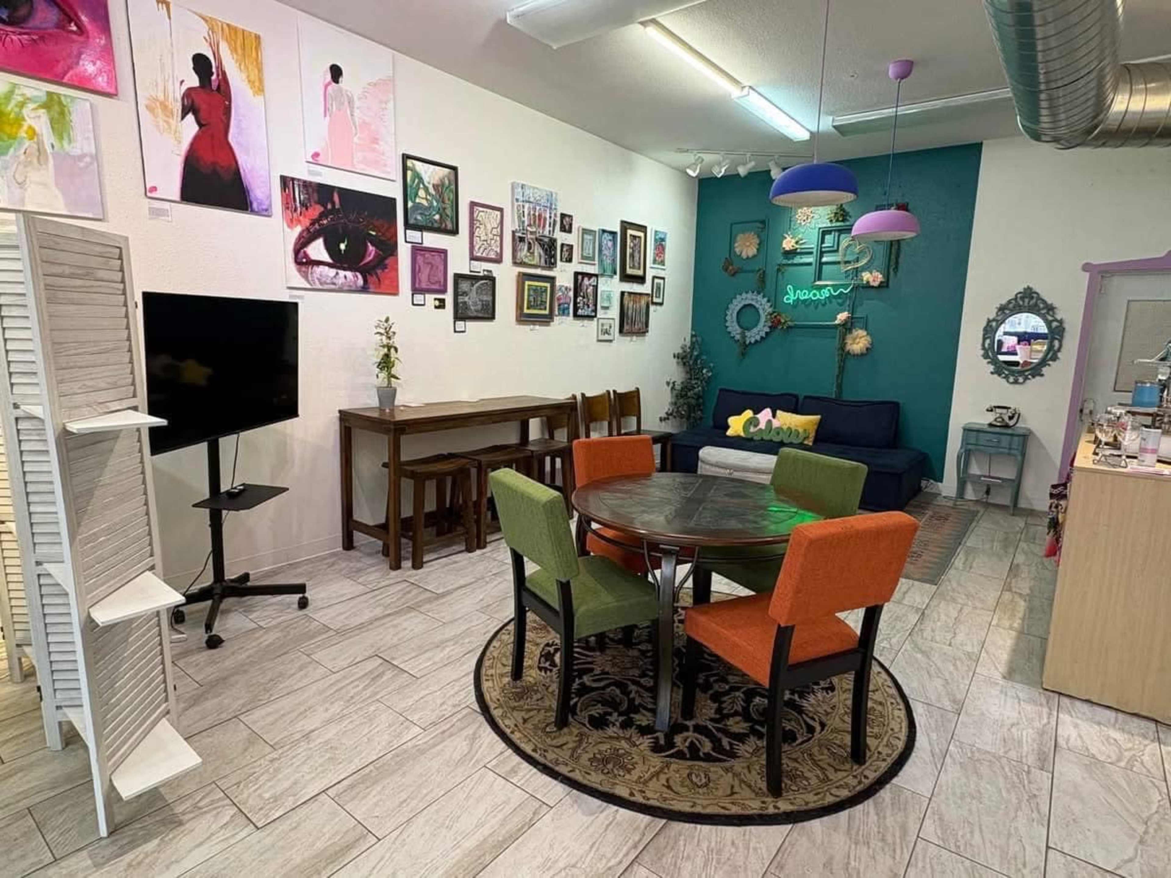 The image shows a cozy interior space featuring a round dining table surrounded by four chairs, a sofa, various artworks on the walls, and a television on a stand.